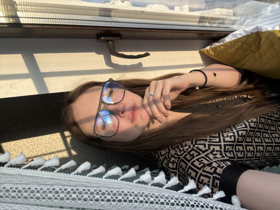 A person with glasses lies on a bed next to a window. The sunlight casts shadows on the bed and the wall. The person wears a patterned shirt and a ring. A yellow pillow is visible behind the person's head. A decorative bedspread with tassels covers the bed. The window has a dark frame and a handle.