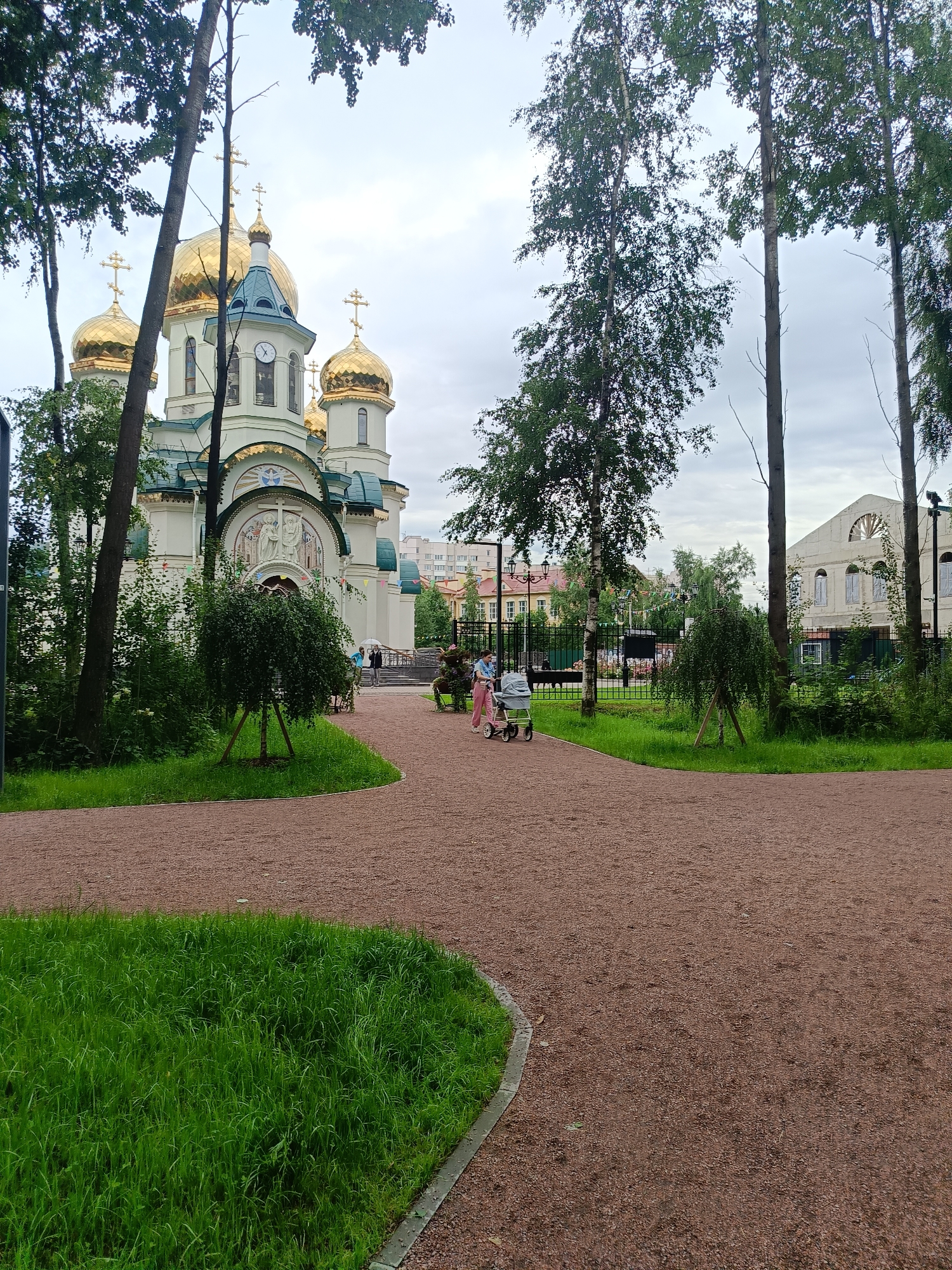 The image depicts the Church of the Resurrection of Christ in St. Petersburg, Russia. The church, with its distinctive golden domes and white facade, is partially obscured by tall trees. A gravel path curves through a grassy area in the foreground, leading towards the church. Two women, one pushing a stroller, are walking on the path in the middle ground. In the background, other buildings and a fence are visible. The sky is overcast.