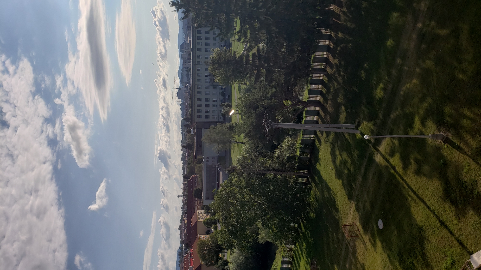 This is an aerial view of a city scene, likely Prešov, Slovakia, featuring a mix of residential buildings, a large school or institutional building, and green spaces. The foreground consists of a grassy area with a single lamppost casting a long shadow, and a small soccer goal in the distance. A metal fence runs along the edge of the green space, separating it from a line of trees. In the midground, a large, multi-story building with many windows dominates the scene, flanked by smaller trees and some parked vehicles. Beyond this, a cluster of older residential buildings with red-tiled roofs is visible. The background is a hazy cityscape with distant hills and a partly cloudy sky. The lighting suggests it is either late afternoon or early morning, with the sun casting distinct shadows.