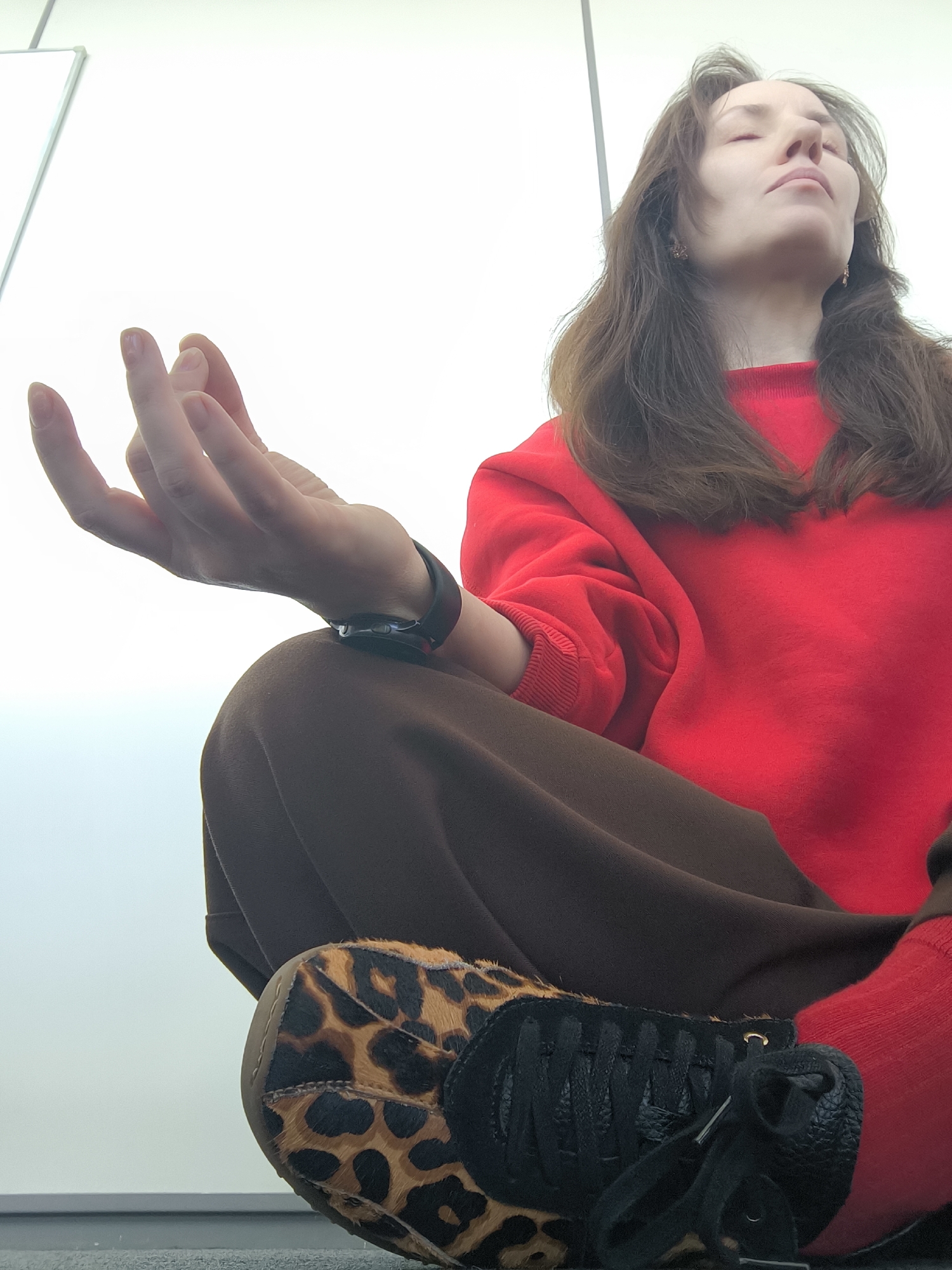 A low-angle medium shot depicts a woman meditating indoors against a white background, likely a wall. She is seated with legs crossed, wearing a red sweatshirt, brown pants, and red socks. On her feet are sneakers with leopard print accents. Her right hand is raised in a meditative pose, and she wears a black smartwatch on her wrist. Her eyes are closed, and her face is tilted upwards. The location is Unknown, Unknown.
