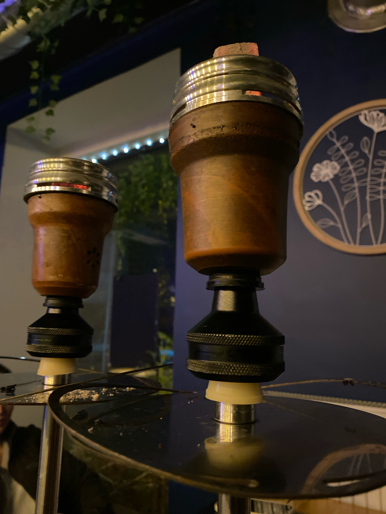 The image depicts two hookah heads, both featuring wooden bowls with metal rims and charcoal pieces on top. The hookah heads are positioned above a reflective dark surface, likely the base of the hookah. In the background, a wall with a circular decorative piece depicting white flowers on a dark blue surface is visible. Green foliage hangs from the ceiling on the left side of the frame, and string lights are illuminated across the background. The setting appears to be an indoor establishment, possibly a lounge or cafe, with ambient lighting. The image does not provide any specific information about the city of Gliwice, Poland, or any individuals present.