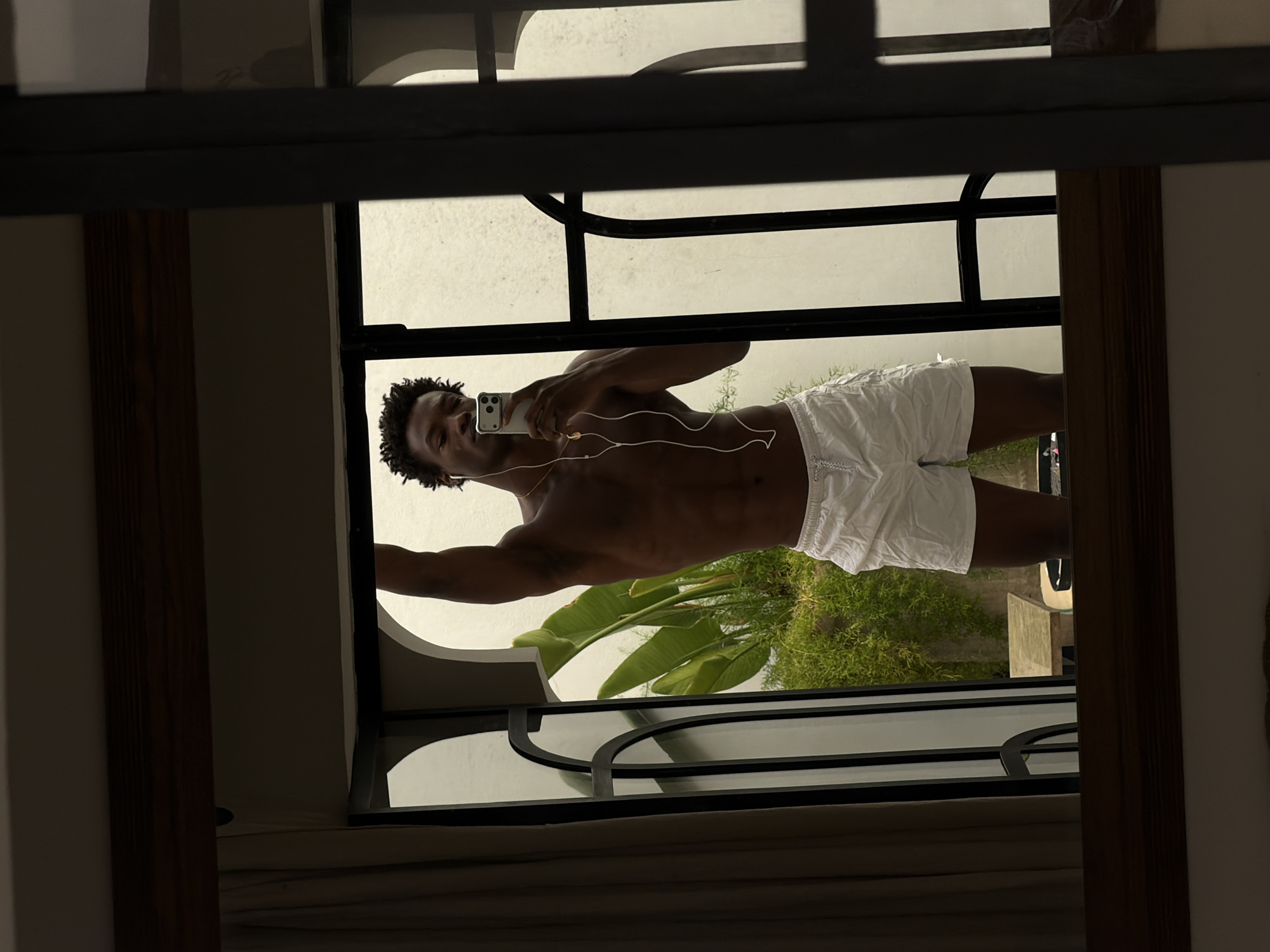 A young Black man with dark, curly hair is taking a mirror selfie indoors. He is shirtless, wearing white swim shorts with a drawstring. He has on white earphones and a thin gold chain with a pendant. He is holding a smartphone in his right hand, angled to capture the reflection. The background is partially visible through a window or doorway, showing lush green plants and a white wall. The setting appears to be a contemporary interior with a dark wood frame around the mirror and architectural elements in the background. The image suggests an indoor space in a warm climate, possibly a hotel or a residential setting in Marrakesh, Morocco, given the presence of tropical plants and the architectural style.