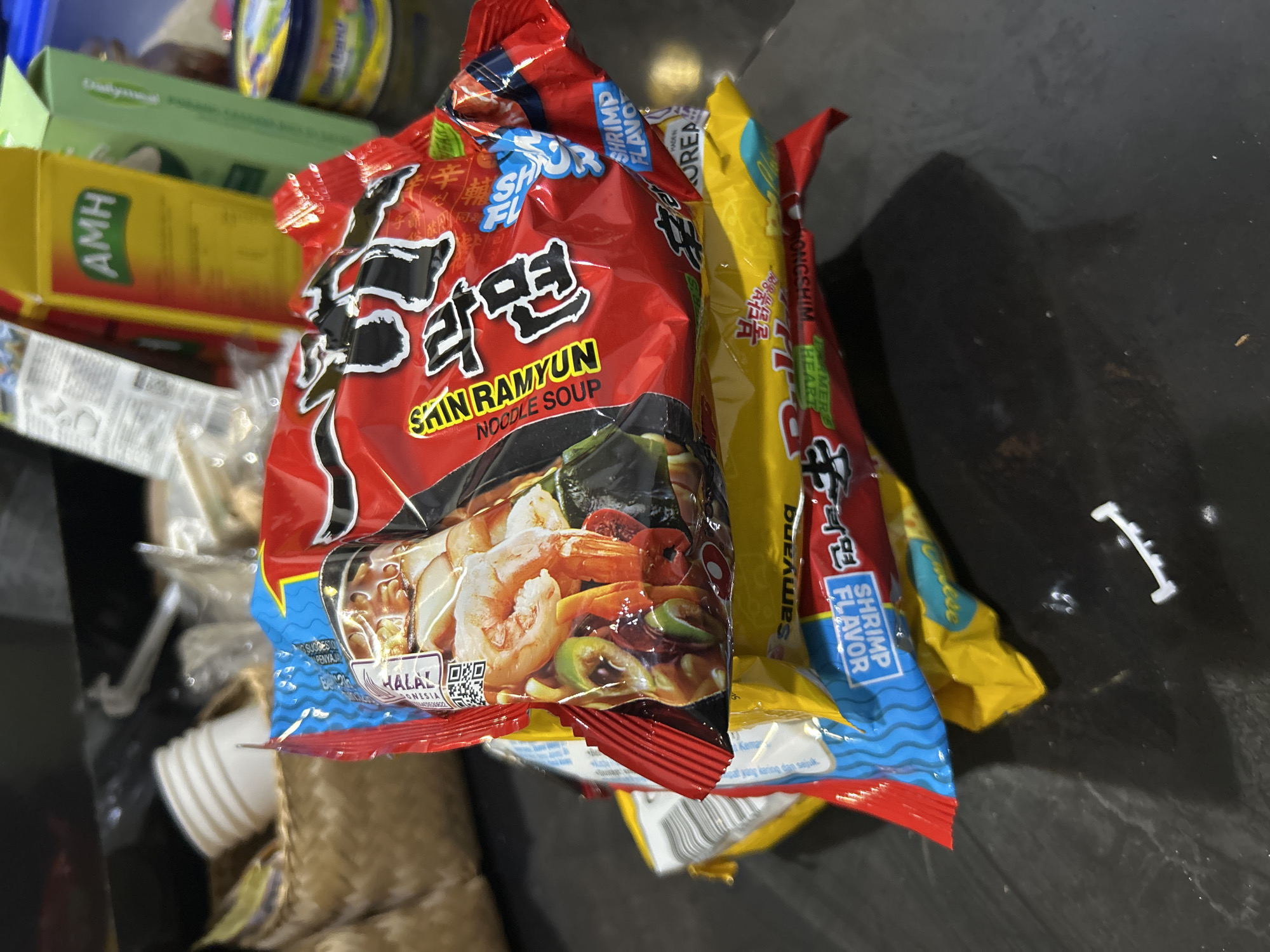 In a close-up, overhead shot, several packets of instant ramen noodles are piled together. The most prominent packet is red and features the brand name "Shin Ramyun" in bold English text, above Korean characters and a depiction of the noodle soup with shrimp. A "Halal" certification is visible on this packet. Adjacent to it is a yellow packet, also appearing to be instant noodles, with blue text that reads "Shrimp Flavor." Other packets are partially visible, including one with red and blue accents. In the background, a yellow box with the letters "AMH" printed on it is stacked with other items, suggesting a retail or storage environment. The scene appears to be indoors, with the lighting suggesting daytime, though no windows are visible. The overall impression is of a casual collection of food items, possibly in a kitchen or pantry setting, with an emphasis on the colorful packaging of the ramen. The visible text "Sukawati, Indonesia" provides a specific geographical context for the image.