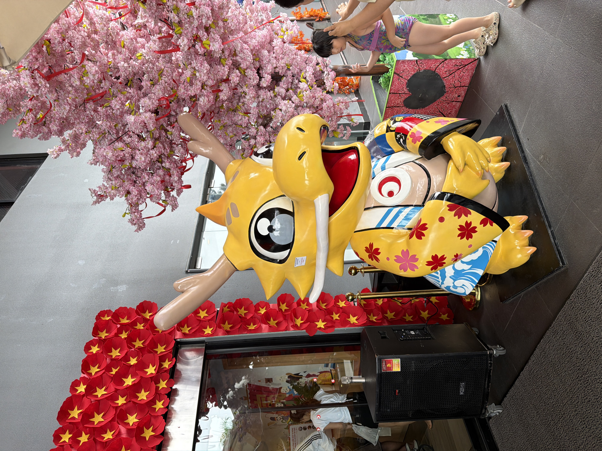 This image captures a vibrant outdoor scene in Da Nang, Vietnam, likely during a festive occasion. A large, bright yellow dragon statue, adorned with floral patterns and an expressive cartoon-like face, is the central focus. To the left of the dragon, a speaker sits on a dark carpeted floor, and behind it, a storefront window reveals people and decorations. A wall decorated with red and yellow starbursts frames the entrance. To the right of the dragon, a cherry blossom tree with pink flowers and red ribbons adds a touch of spring and celebration. A young girl in a swimsuit stands near the tree, interacting with someone whose hands are visible at the edge of the frame. The overall atmosphere is festive and visually engaging, hinting at a public event or decoration.