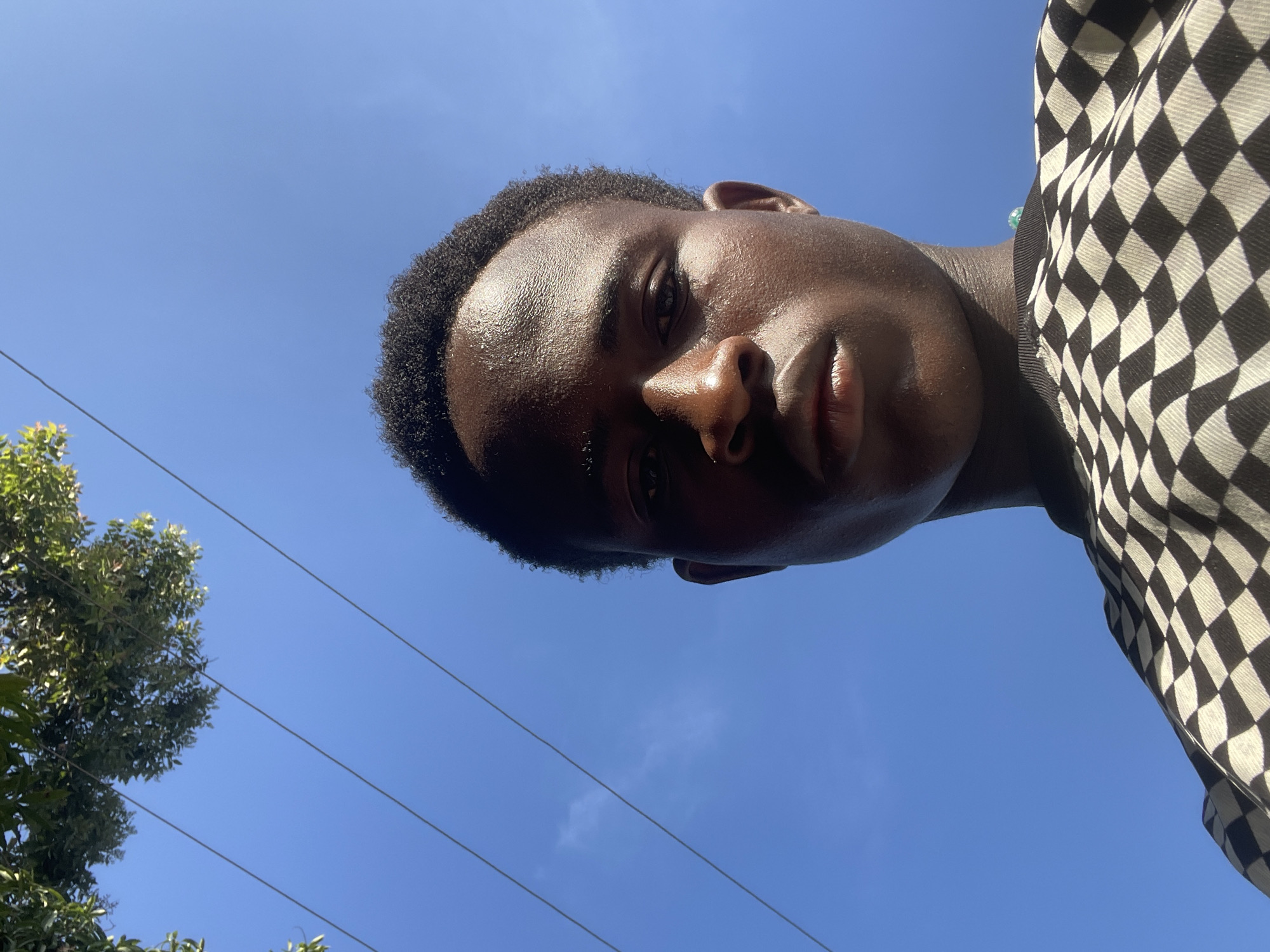 A close-up, low-angle shot shows a young Black male looking directly at the camera. He has dark skin and short, textured hair. His face is slightly shiny, possibly from sweat or oil. He is wearing a black and white checkered t-shirt. The background consists of a clear blue sky with a few wispy clouds. Overhead, green tree leaves and power lines are visible. The overall lighting suggests it is daytime. The image is a selfie, taken outdoors in what appears to be an urban or suburban setting. The image is not sufficient to determine the specific location within Jos, Nigeria, beyond it being an outdoor, daytime setting with vegetation and power infrastructure.