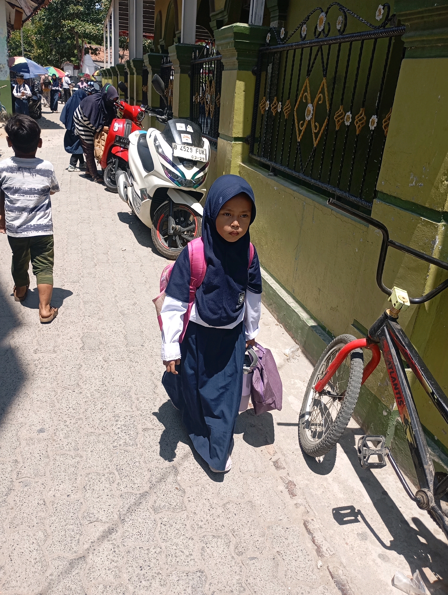 In the city of Babelan, Indonesia, on a sunlit day, a young girl wearing a navy blue hijab and school uniform walks down a paved street. She carries a light purple bag in her right hand and has a matching backpack on her shoulders. To her left, a boy in a striped t-shirt and green pants walks away from the camera. In the background, several people are seen walking, some with colorful umbrellas, near parked motorcycles. A black and red bicycle leans against a light green wall adorned with a decorative metal fence. The scene depicts a typical day in a residential or school area.