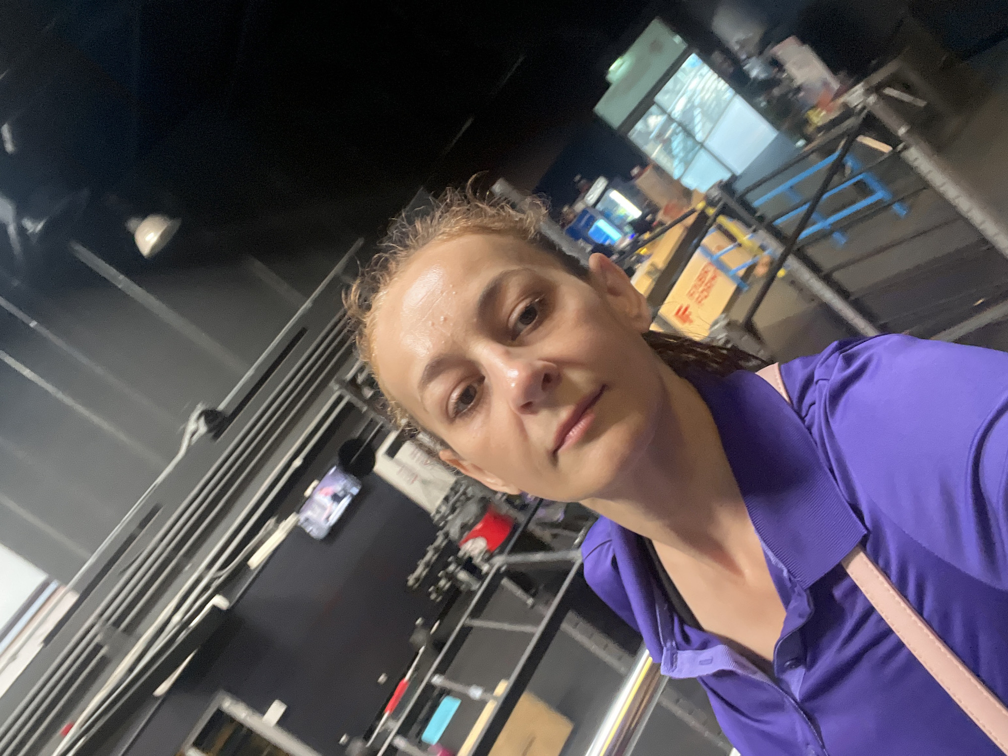 A woman with blonde hair pulled back is taking a selfie in what appears to be a gym or fitness facility. She is wearing a purple polo shirt and a light pink crossbody bag. The background shows gym equipment, metal racks, and what looks like a television screen mounted on the wall. The lighting is somewhat dim, and the overall impression is of a casual, in-the-moment snapshot. The exact location within the city is unknown.