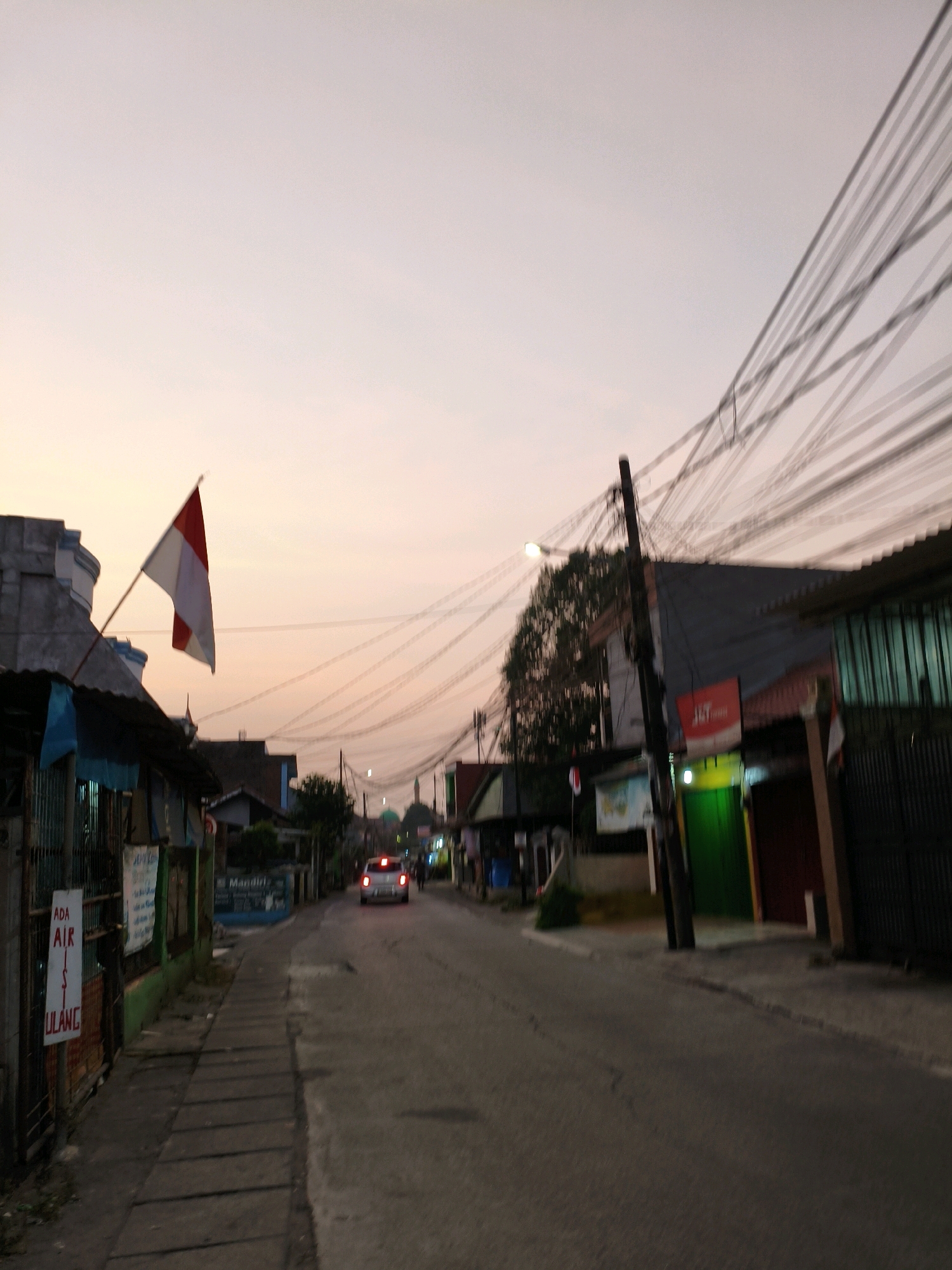 The image depicts a street scene at dusk in Bekasi, Indonesia. The sky exhibits a soft, orange-pink gradient, indicative of sunset. A red and white Indonesian flag is prominently displayed, attached to a building on the left side of the street. Adjacent to the flag, a row of low-rise buildings lines the street, some with corrugated metal roofs. A sidewalk runs along the left side of the street, marked by concrete slabs. On the right side, more buildings are visible, including one with a green door and another with a red and brown facade. A prominent utility pole with a dense network of wires crosses the upper portion of the image. A white car with illuminated taillights is driving away from the camera down the asphalt road. In the background, the silhouette of a mosque with a green dome is discernible. Signs are present on some of the buildings, though they are largely illegible due to image quality and distance. The overall impression is that of a typical residential or commercial street in a Southeast Asian urban environment during the transition from day to night.