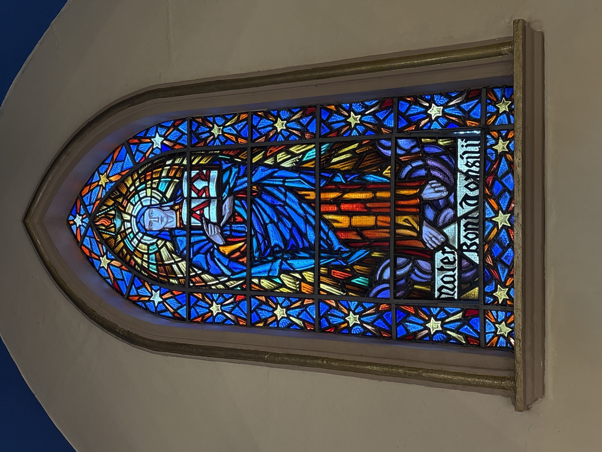 A stained glass window depicts a figure, likely the Virgin Mary, dressed in a blue robe. She holds an open book with an inscription that reads "Mater Boni Consilii". The figure is surrounded by a background of blue and yellow glass fragments shaped like stars and other geometric patterns. The window is framed by an arched stone structure with a cream-colored wall on either side. The window is located in the São Paulo Cathedral (Catedral da Sé) in the city of São Paulo, Brazil.