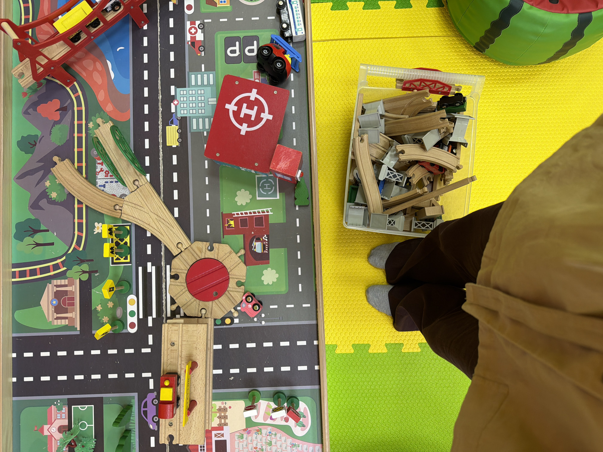 The image shows a child's play area on a yellow and green foam tile floor. On the left side of the image, there is a wooden toy railway track and town play mat with roads, buildings, and scenery printed on it. The railway track is made of light brown wood and includes a red turntable. Several wooden toy cars and a bridge are visible. A red wooden building with a white "H" on top is also present. To the right of the play mat, there is a clear plastic container filled with wooden railway tracks. A person wearing brown pants and gray socks is standing next to the container. A green watermelon-shaped object is visible in the upper right corner. The location cannot be determined from the image.