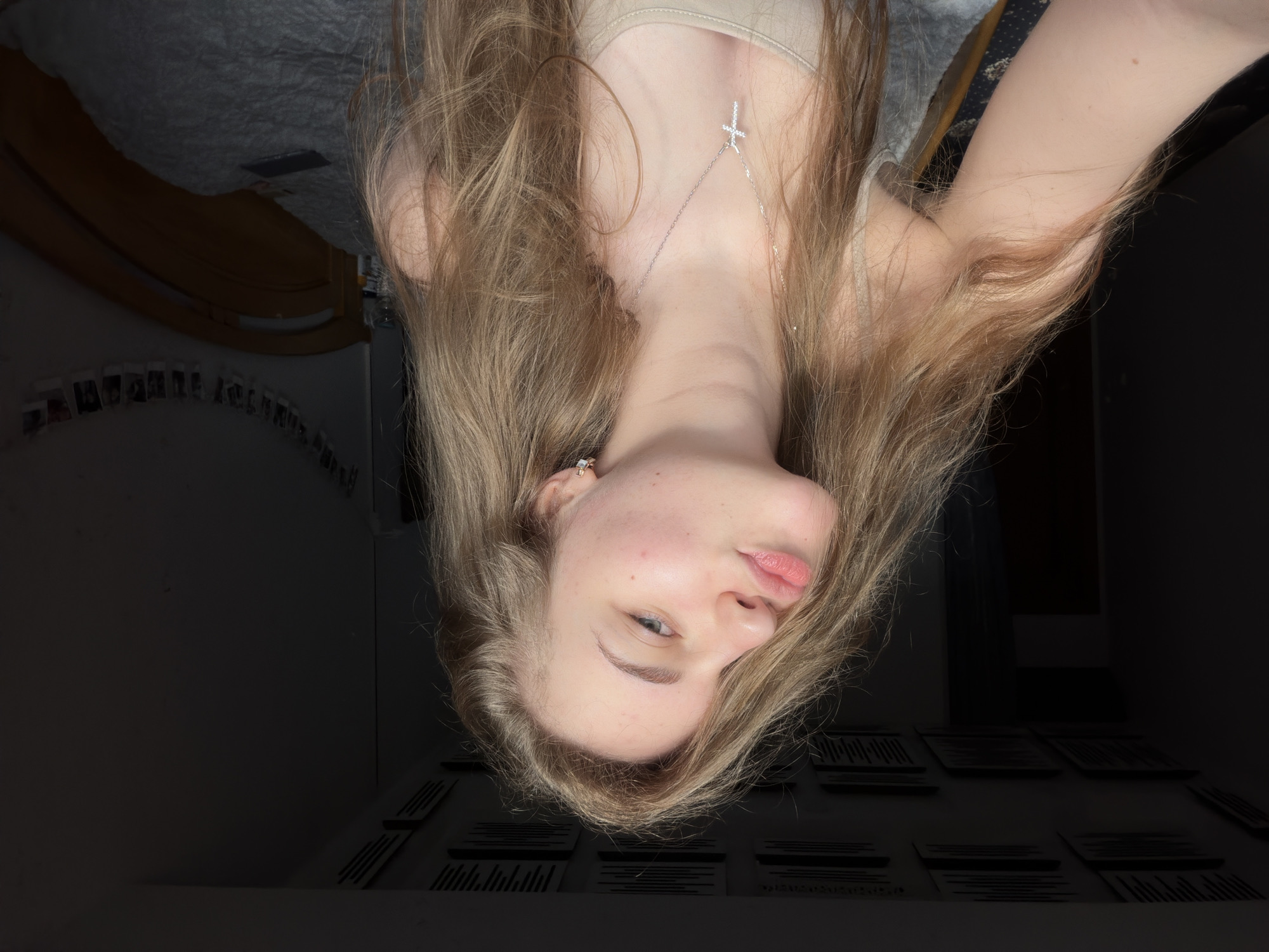 A young woman with long, blonde, wavy hair is captured in a selfie. She is wearing a beige tank top and a silver necklace with a cross pendant. Her hair is falling across her face, obscuring part of her left side. She has on small, sparkling earrings. Her lips are pursed in a "duck face" pose, and her right eye is looking towards the camera. The background suggests an indoor setting, possibly a bedroom, with a wooden bed frame and a string of photos hanging on the wall. The lighting is dim, creating shadows. The image does not provide any specific details to ascertain the location within the city of Dnipro, Ukraine.