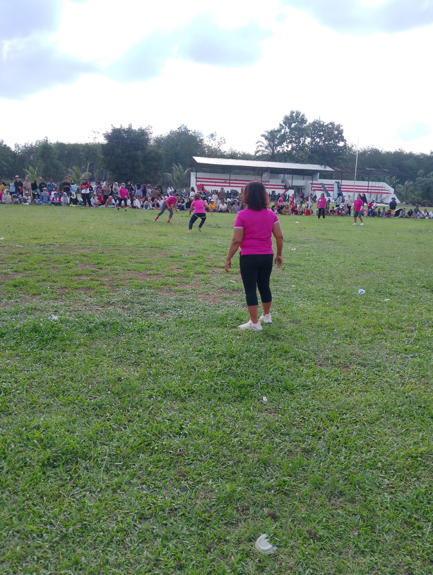 In Serbelawan, Indonesia, a group of people are gathered on a grassy field, seemingly for an outdoor event. A woman in a pink t-shirt and black pants stands in the foreground, facing away from the camera. In the background, several individuals in pink shirts are engaged in what appears to be a sporting activity on the field. Spectators are seated and standing along the edge of the field, with a building structure and trees visible behind them. The sky is overcast with scattered clouds.