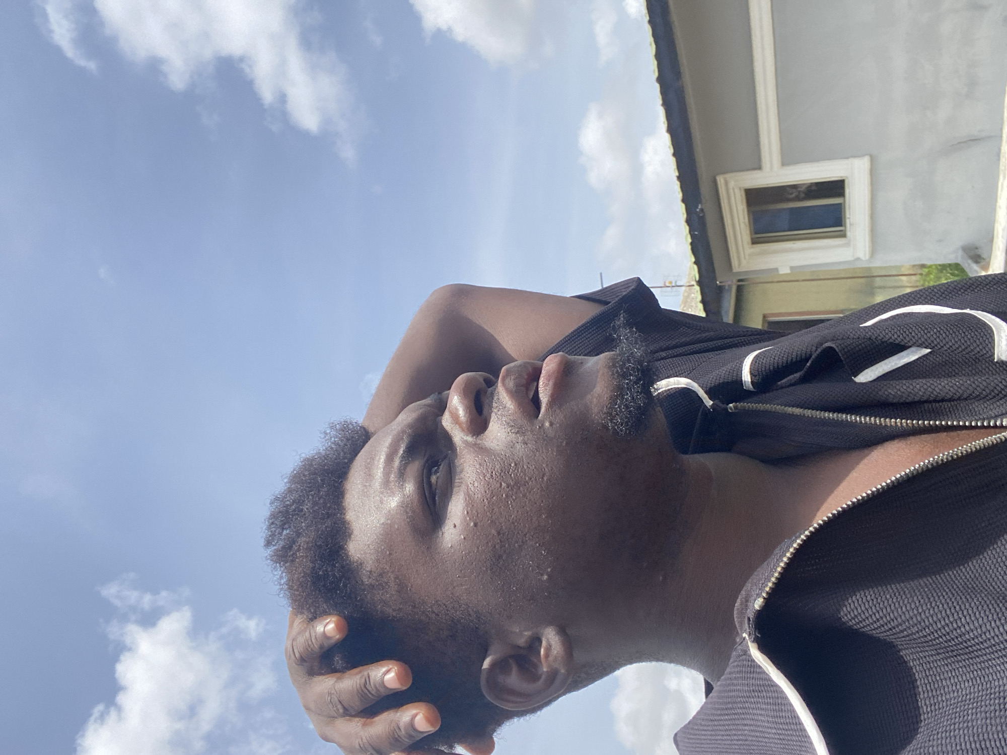 A low-angle shot shows a Black man with short, curly hair and a light mustache, looking up with his hand on his head. He is wearing a black zip-up jacket with white stripes. Behind him, the sky is light blue with scattered white clouds. In the upper right corner of the frame, a portion of a light gray building with a window is visible. The image appears to be taken outdoors during the daytime in Unknown, Unknown.
