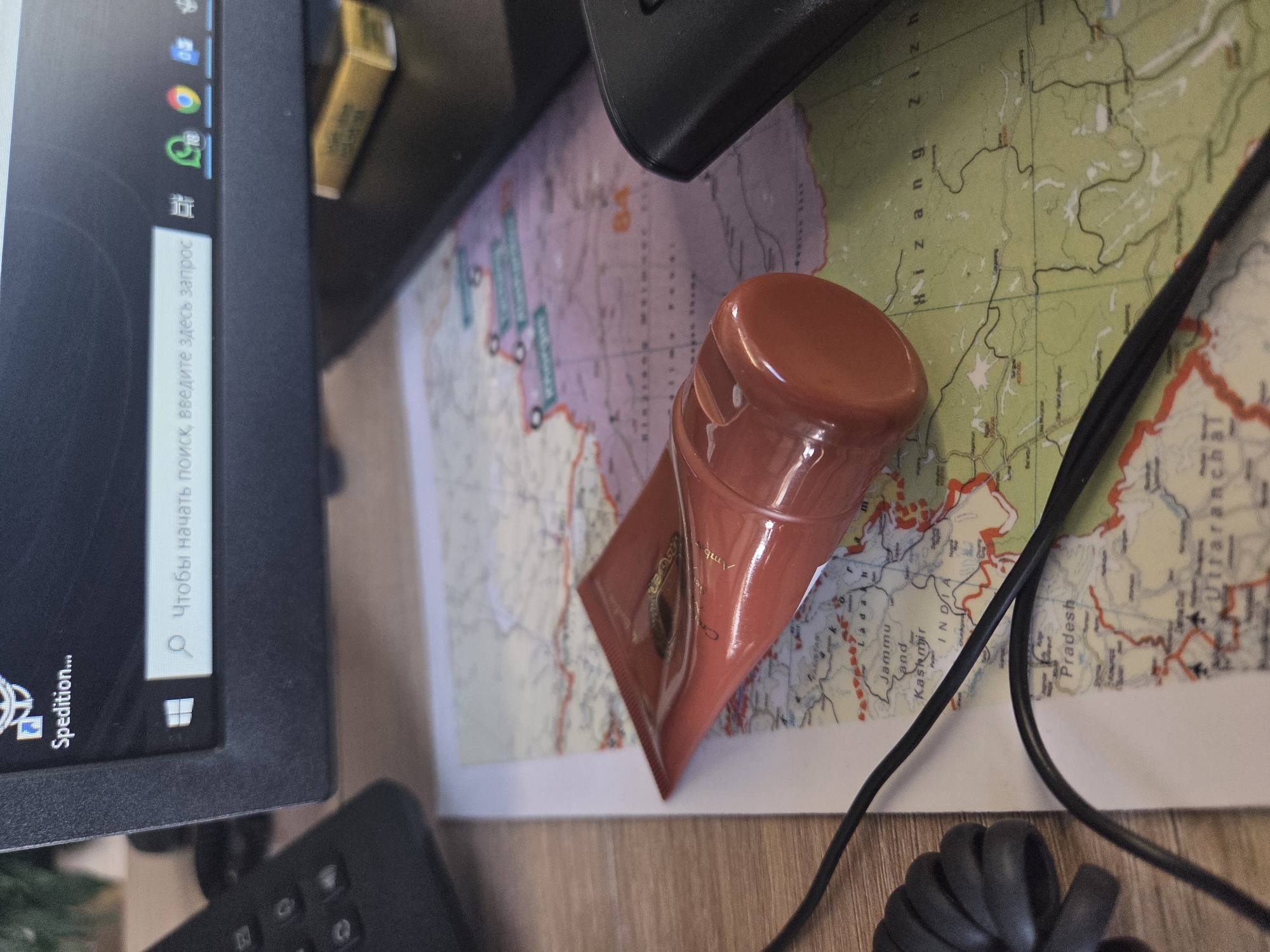 The image shows a desk scene with a computer monitor on the left, displaying a search bar with the text "Чтобы начать поиск, введите здесь запрос". A brown tube is centrally placed on a map showing Jammu and Kashmir, India, with the text "INDIA Jammu and Kashmir" visible. A black computer cable is also present on the map, coiled at the bottom. The environment appears to be a workspace, with the map covering a part of the desk surface.