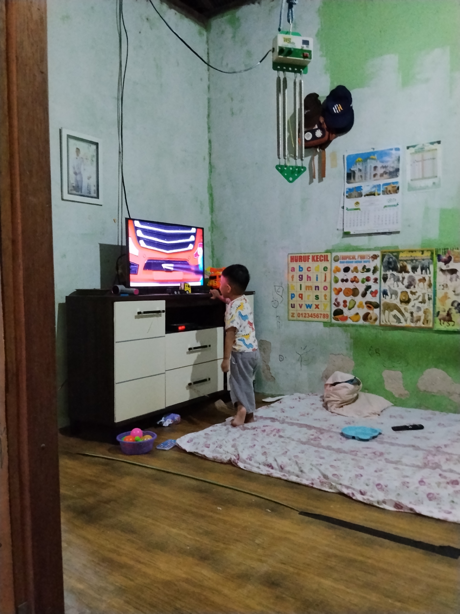 In Bumi Etam, Indonesia, a young boy watches television in a sparsely furnished room. The room has pale green walls, with some peeling paint and water stains. A wooden dresser with a dark top and cream-colored drawers stands against the wall, holding a television displaying a cartoon. A small toy truck is visible on the dresser. The boy, wearing a colorful t-shirt with dinosaur prints and gray pants, stands facing the television. To the right of the dresser, a mattress covered with a floral sheet lies on the floor, with a pillow and a remote control. Educational posters with alphabets and pictures of animals and fruits are hung on the wall. A framed photograph of a couple in formal attire hangs on the wall to the left. A small basket with colorful balls is on the wooden floor near the dresser. Various wires are visible running along the ceiling and walls. The overall impression is of a simple, lived-in space.