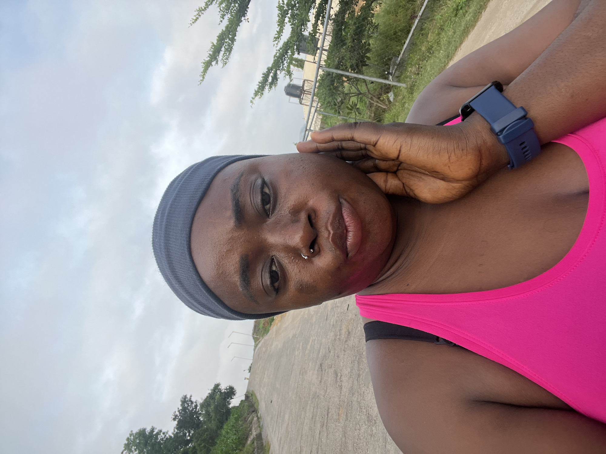A dark-skinned woman with a grey headband and a pink athletic top is looking into the camera, holding her right hand to her cheek. She has a nose ring. She is wearing a blue smartwatch on her left wrist. The background shows a paved road curving uphill, with streetlights visible in the distance. Trees and a chain-link fence are present on the right side of the frame. Two water tanks on elevated platforms are visible behind the fence. The sky is overcast. The image appears to be taken in Abuja, Nigeria.