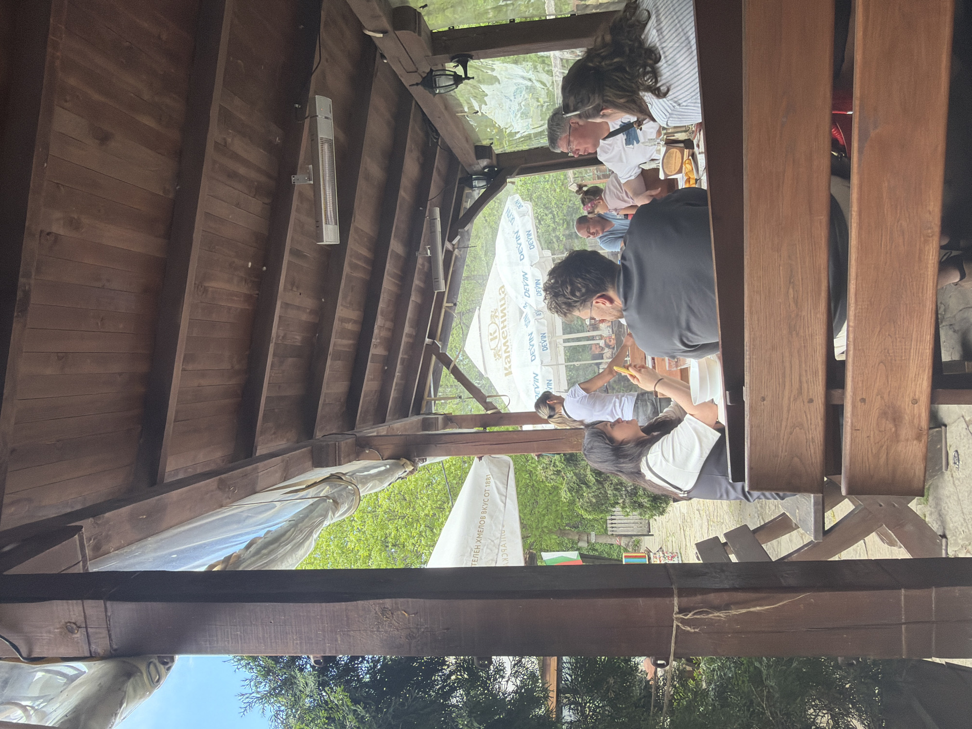 Here's a forensic description of the image:
**Scene:** An outdoor dining area, likely a restaurant or cafe, located in what appears to be Dryanovo, Bulgaria, based on the Bulgarian flag visible. The dining area is covered by a wooden structure.
**People:** Several people are seated at tables. The focus is on one table where several individuals, men and women, are seated and engaged in eating or conversation. One person is holding what appears to be food or a phone.
**Objects:** The scene contains wooden beams, tables, chairs, umbrellas bearing the brand "Devin", a heating lamp, and other restaurant paraphernalia.
**Actions/Interactions:** The group of people is dining together, with some individuals interacting with food or perhaps using their phones.
**Environment:** The setting is outdoors with trees and greenery visible beyond the dining area. The lighting suggests it is daytime.