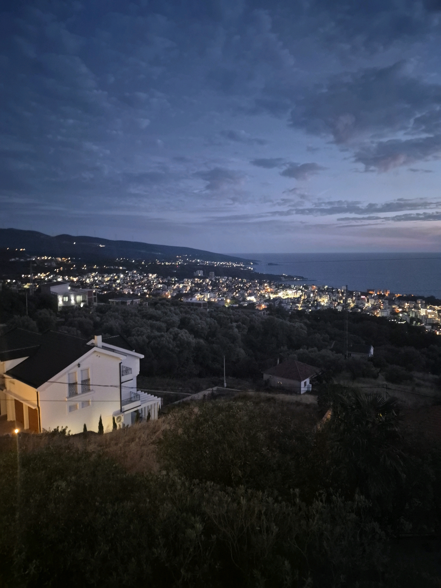 The image depicts a twilight scene overlooking the coastal city of Dobra Voda, Montenegro. In the foreground, a two-story white house with a black roof is partially visible, with balconies and white railings. Several trees and dense shrubbery surround the house. Beyond the immediate foreground, a vast expanse of olive trees covers a hillside, leading down to the brightly lit city that sprawls along the coastline. The city is a dense collection of buildings with numerous illuminated windows, suggesting a populated urban area. The Adriatic Sea forms the backdrop, stretching to the horizon under a cloudy, deep blue sky. The lighting suggests it is either dusk or early dawn, with artificial lights from the city being prominent. The overall impression is of a serene, yet vibrant, coastal settlement at the transition between day and night.