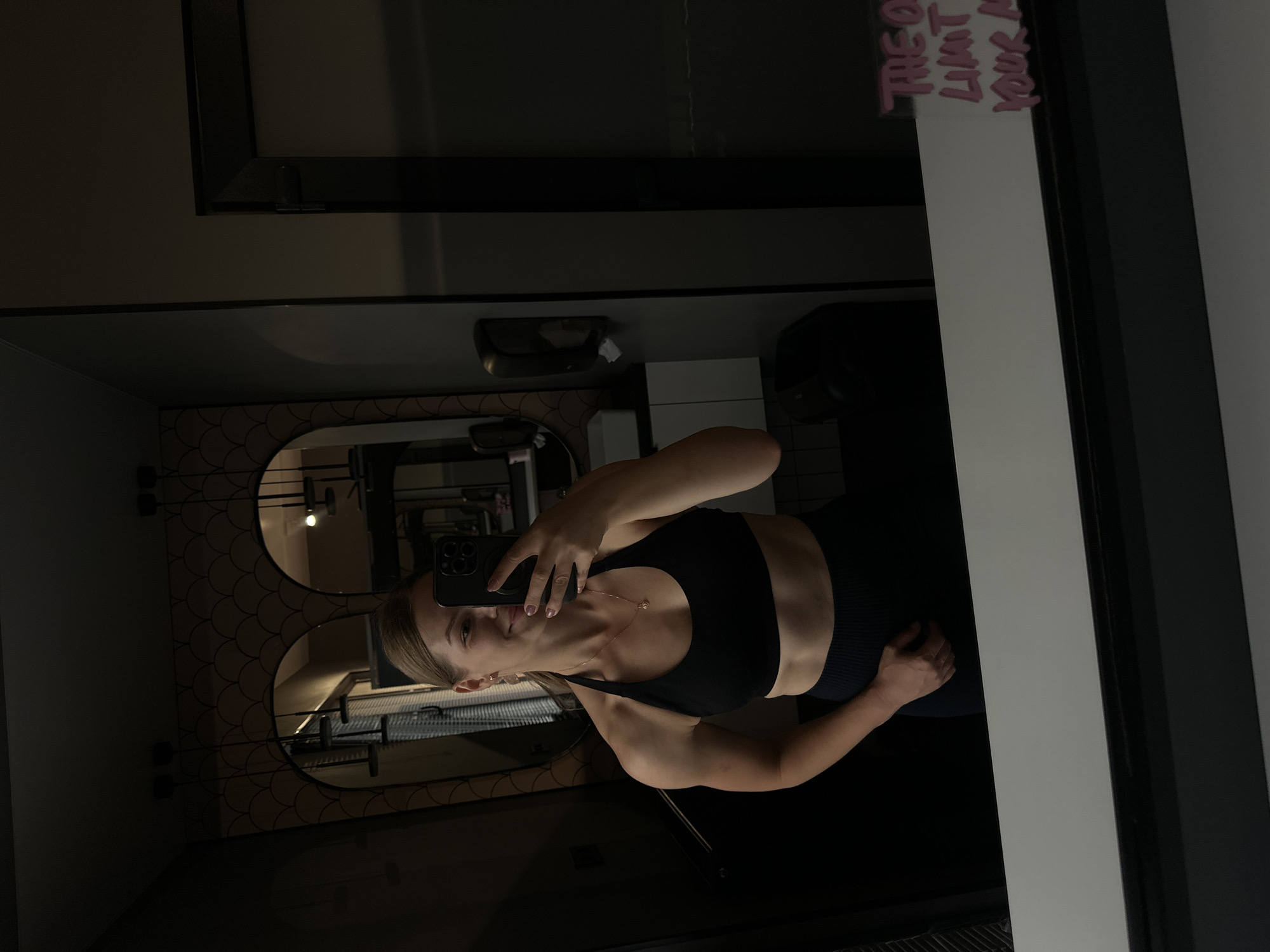 A woman with blonde hair pulled back into a ponytail, wearing a black sports bra and black leggings, is taking a mirror selfie in what appears to be a modern bathroom. She is smiling and holding a black smartphone in her right hand. Her left arm is bent at the elbow and her hand is resting on her hip. The bathroom features a large mirror, ornate wall sconces, and a wall-mounted dispenser. In the reflection, a towel rack and a toilet can be partially seen. On the lower right side of the mirror, pink handwritten text reads "THE ONLY LIMIT IS YOUR MIND". The image does not contain information that allows for the specific location in the city of Ternopil, Ukraine to be identified.