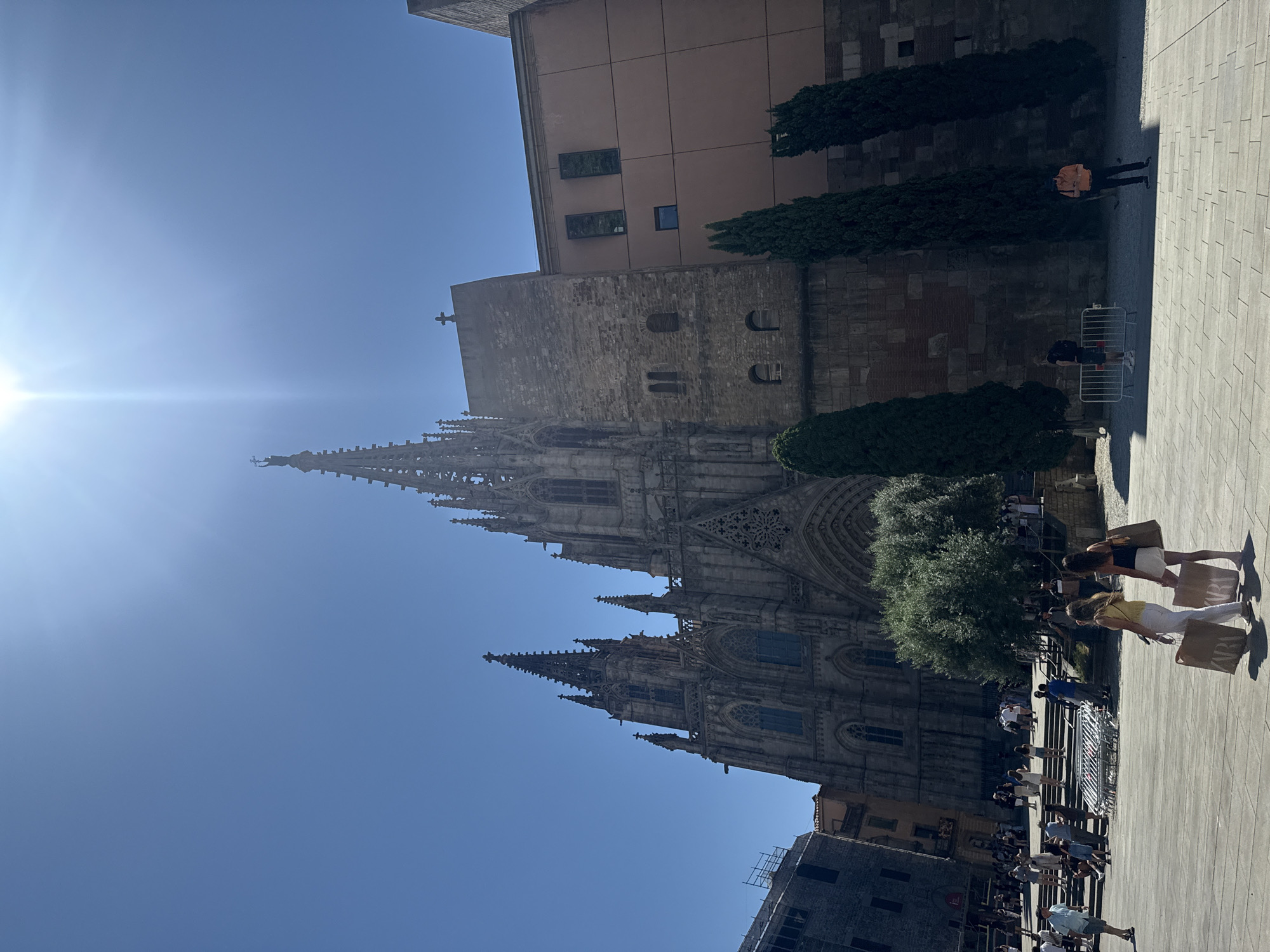 This image shows the Barcelona Cathedral on a bright, sunny day. The Gothic cathedral, a prominent landmark in the Gothic Quarter of Barcelona, Spain, is viewed from a public square. The architecture is characterized by its spires, ornate facade, and large arched windows. In the foreground, people are gathered in the square, some walking and others sitting. Two women in the mid-ground are carrying shopping bags. The square is paved with large, light-colored tiles. Tall, slender cypress trees are planted along the side of the cathedral. The sky is a clear, vibrant blue, with the sun creating lens flare effects in the upper portion of the image. The scene captures a bustling urban environment with a focus on the historical cathedral.