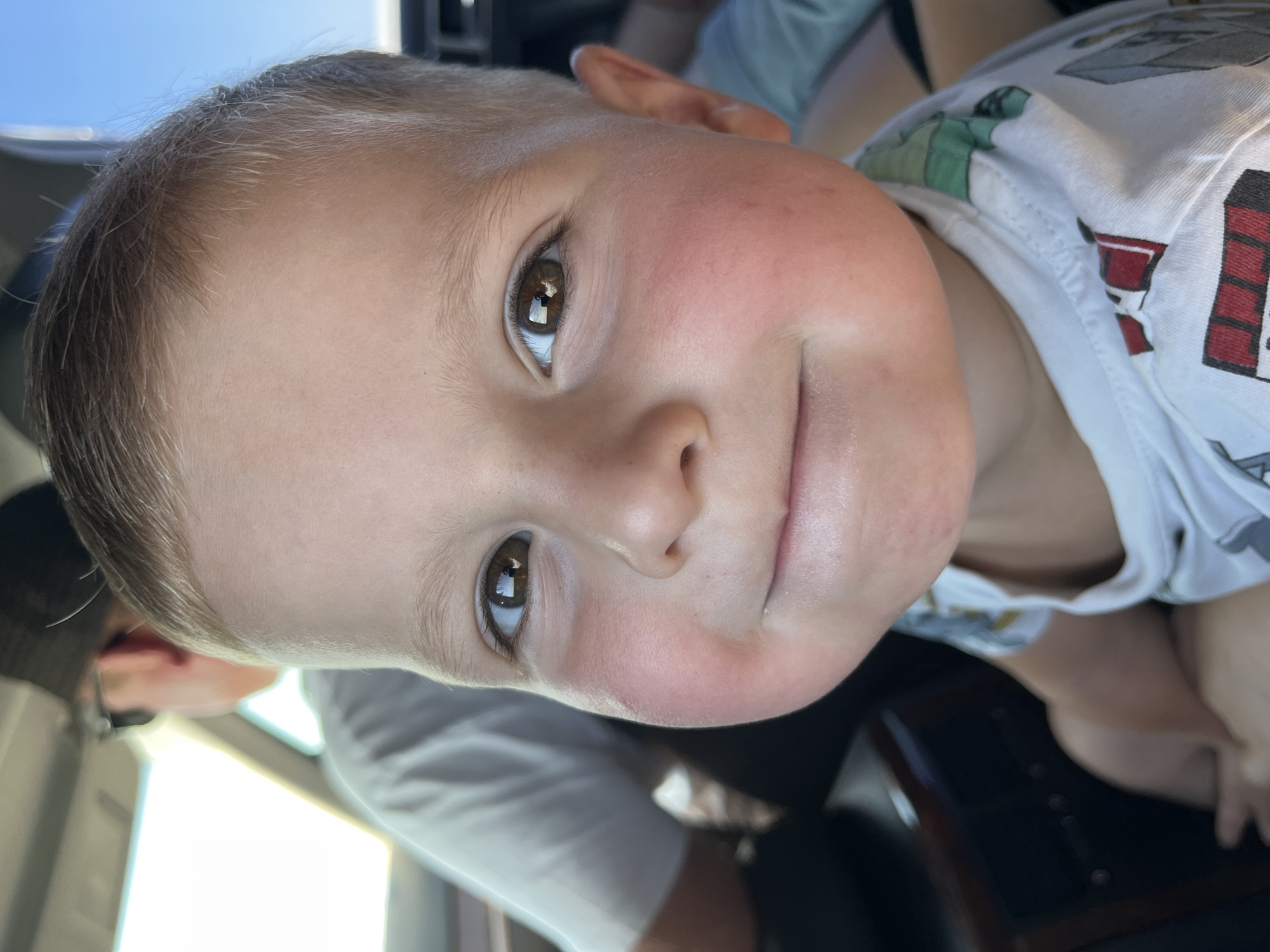 A close-up, eye-level shot shows a young boy with fair hair and brown eyes, looking directly at the camera. His cheeks are flushed pink, and he has a slight smile. He is wearing a white t-shirt with a colorful pattern. In the background, out of focus, a person wearing a black baseball cap and sunglasses is visible, appearing to be in the driver's seat of a vehicle. The scene suggests an indoor setting, possibly within a car, as indicated by the presence of a person in the driver's seat and what appears to be a car interior. The lighting suggests it is daytime. The location of Shumeika, Russia, cannot be determined from the visual information provided in the image.