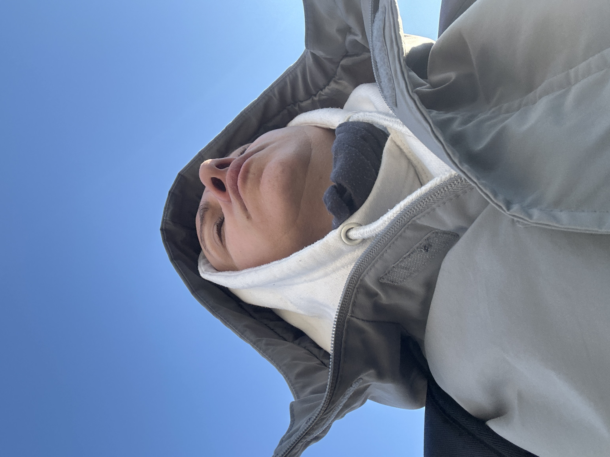 A low-angle shot captures a person against a clear blue sky. The person is wearing a white hooded sweatshirt under a gray jacket with the hood up. A dark gray or black object is visible inside the white hoodie, near the person's cheek. The person's face is visible, with a focus on their nose, lips, and eye. There is no discernible information to ascertain whether the location is in the city of Sofia, Bulgaria.