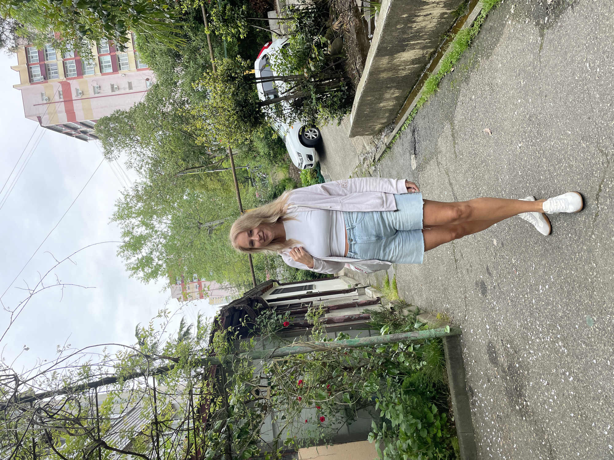 A woman stands on a gray, cracked asphalt road, near a sidewalk curb to the right and green shrubbery to the left. She is wearing white sneakers, light blue denim shorts, a white top, and a pale pink jacket. Behind her is a weathered, overgrown house with a dark roof and faded paint, partially obscured by climbing plants. Further back, a white car is parked amidst trees and greenery. A multi-story apartment building with a light pink and beige facade stands in the background under a cloudy sky. Based on the architecture, vegetation, and overall environment, this scene is likely in Sochi, Russia.