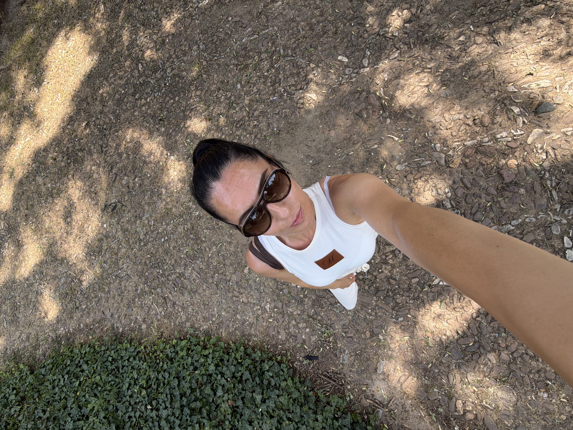A high-angle shot looks down at a woman standing on a gravel path with scattered leaves and rocks, bordered by dense green ivy. The woman, with her dark hair pulled back in a bun, wears sunglasses and a white tank top with a brown square logo on the left chest. Her right arm extends towards the camera, holding it for a selfie. The ground is dappled with sunlight and shadows, suggesting it is daytime. The setting appears to be an outdoor natural area, possibly a park or garden. The woman's expression is neutral. There is no visible text other than the logo on her shirt, which is not legible.