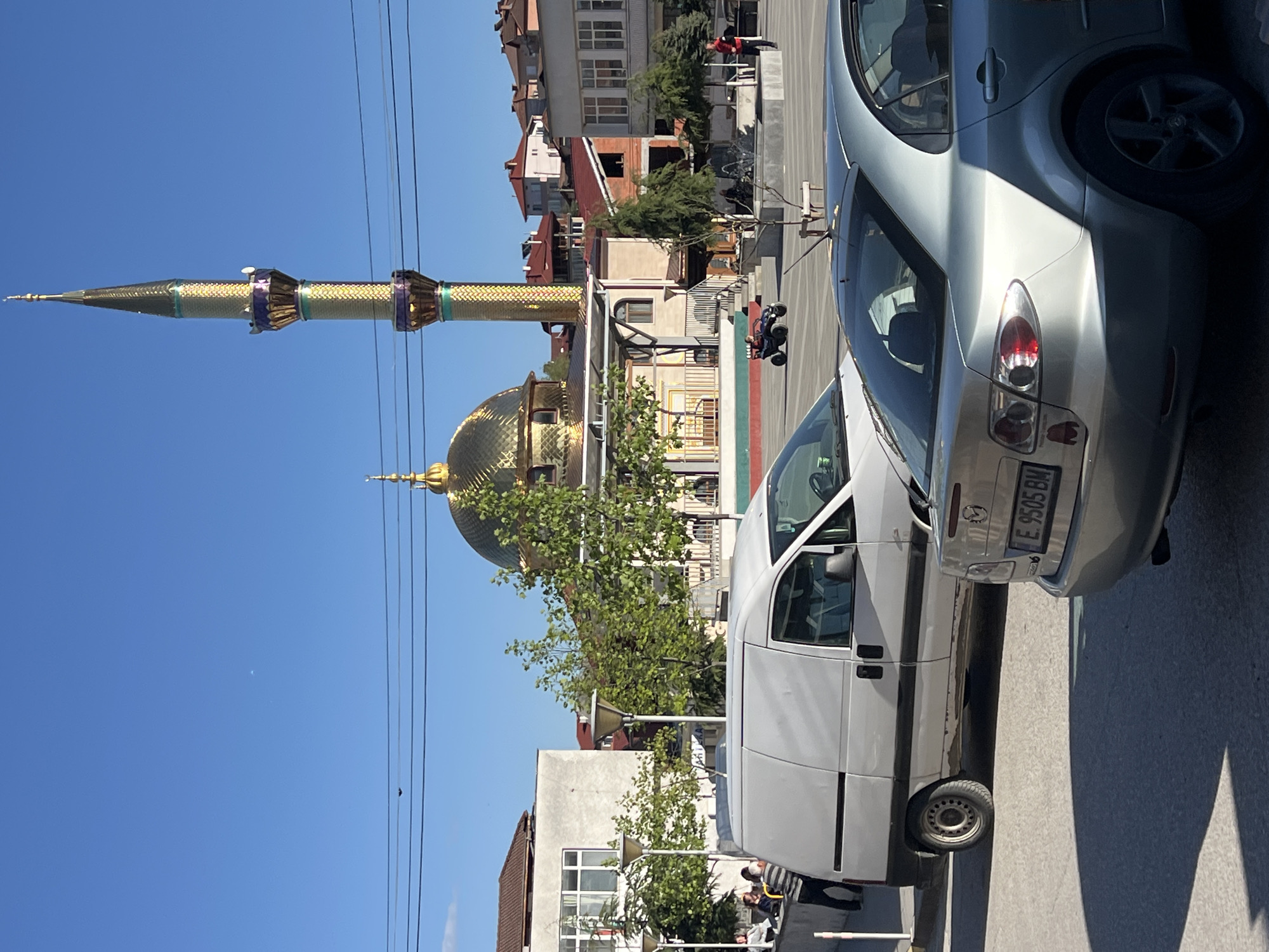 The image depicts a street scene in Breznitsa, Bulgaria, dominated by the Golden Mosque, its golden dome and minaret towering over the cityscape. A white van and a silver car are parked along the street, partially obscuring the view of the mosque. Power lines run across the clear blue sky above. In the background, buildings of varying heights and colors are visible, along with trees and a few people near a railing. A child is seen riding a small vehicle on the street. The scene appears to be captured on a sunny day. The vehicle license plate number is E 9505 BM.