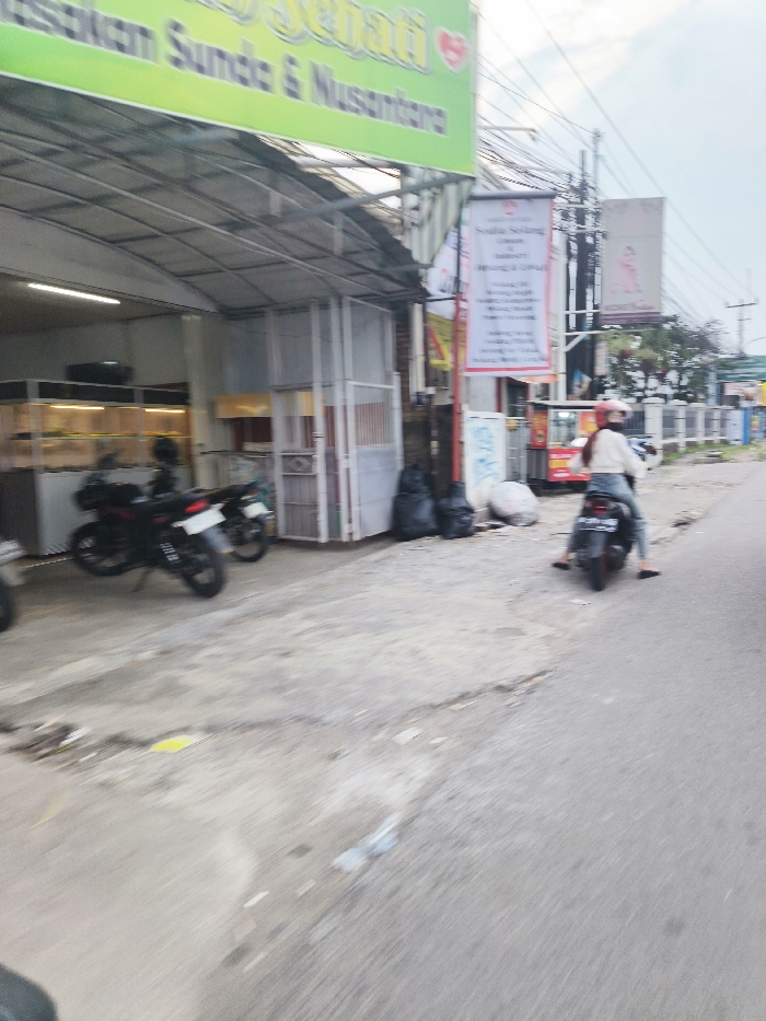 The image shows a street scene in Cileunyi, Indonesia, located at coordinates -6.938 latitude and 107.734 longitude. A woman wearing a white shirt and a red helmet is riding a motorcycle down a street, and there are two parked motorcycles on the side of the road. Behind the motorcycles, a building has a green awning and various signs on the side of the building. The street appears to be paved with concrete, and the road is lined with buildings and trees. There are also some black garbage bags on the side of the road. The image appears to have been taken on a cloudy day.