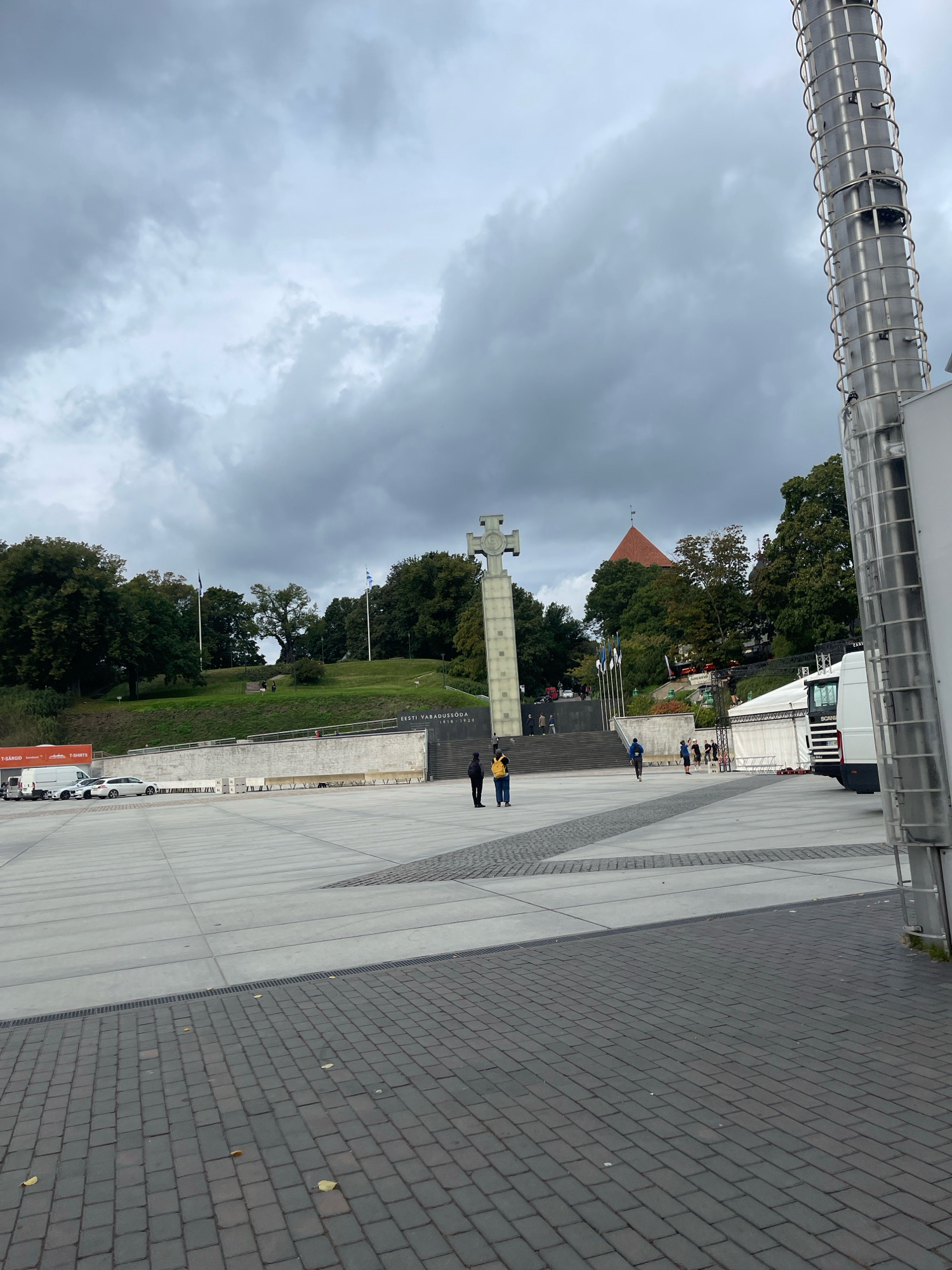 The image depicts a wide, paved plaza in front of a monument with a large cross. The monument is situated on a grassy hill with trees in the background. The sky is overcast with dark, heavy clouds. In the foreground, the plaza is paved with large, grey rectangular tiles, transitioning to dark cobblestones on the right side. A tall, cylindrical metal structure, possibly a light pole or part of a stage setup, is prominently visible on the far right, extending upwards out of frame.
To the left of the monument, a white van and several cars are parked. A white tent structure is visible behind the cars, with a white truck parked nearby. Several people are scattered across the plaza. Two individuals stand near the base of the monument, one wearing a black jacket and the other a yellow backpack. Further back, several other individuals are walking up the steps towards the monument. Flags are visible on poles on the grassy hill.
The monument itself is a tall, concrete structure with a large, stylized cross at the top. Below the cross, there is an inscription that appears to read "EESTI VABADUSSÕDA 1918-1920" which translates to "Estonian War of Independence 1918-1920". This indicates the location is likely the Tallinn War of Independence Victory Column, located in Freedom Square (Vabaduse väljak) in Tallinn, Estonia.
The overall atmosphere is somber due to the overcast sky and the monumental nature of the scene. The presence of a tent and truck suggests a possible event or setup taking place in the plaza.