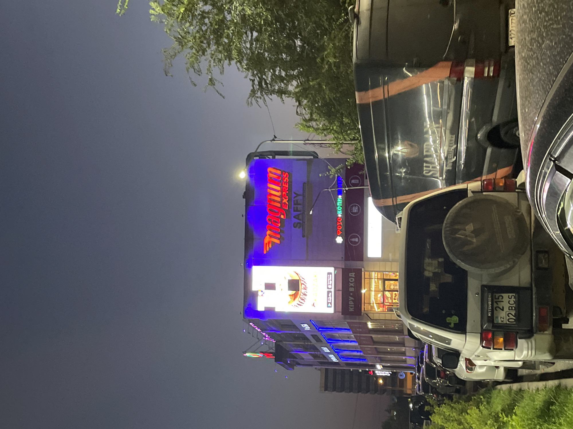 The image captures a street scene in Almaty, Kazakhstan at dusk or night. A prominent building with illuminated signs is visible, featuring the "Magnum Express" logo in bright red and blue lettering, along with other signage in Cyrillic. The building appears to be a commercial establishment, possibly a supermarket or shopping center, with storefronts and entrances visible on the ground floor.
Several vehicles are parked or driving on the street. In the foreground, a white Mitsubishi Pajero SUV is parked, displaying a license plate that reads "215 02 BCS" with "KZ" indicating Kazakhstan. To the right of the SUV, a dark-colored van with orange stripes and the logo "SHAFRAN" is parked. The van has a website address printed on its side, "WWW.SHAFRAN.KZ". Other cars are visible lined up along the street in the background.
Green trees and foliage are present on the right side of the image, partially obscuring the building and the sky. The sky is a deep twilight blue. The overall atmosphere suggests an urban environment with commercial activity at night.