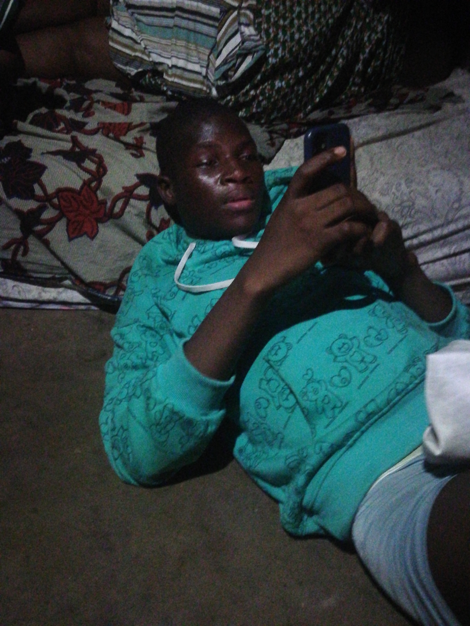 A person lies on a bed using a mobile phone. The bed has two different patterned blankets. The person is wearing a blue hoodie with cartoon bear designs and grey underwear. The floor is brown and appears to be carpeted. The photo was taken indoors at night. The location is unknown; Lagos, Nigeria is not evident.