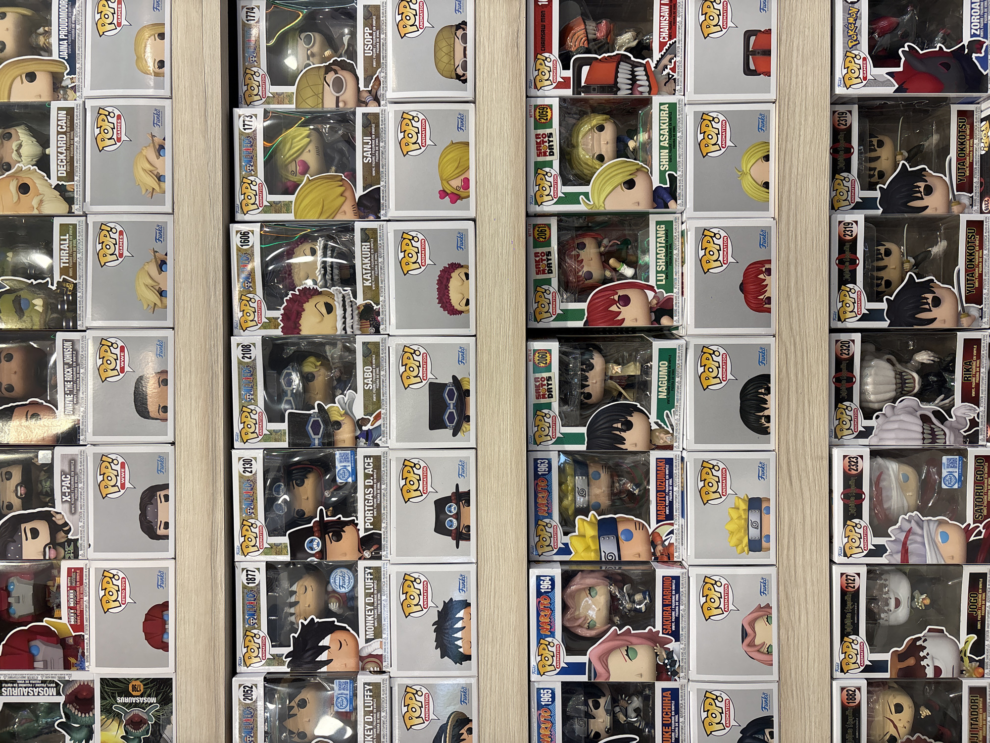 The image shows a collection of Funko Pop! figures, organized and arranged in rows, likely displayed on a shelf. The figures span various franchises including anime ("One Piece," "Naruto," "Jujutsu Kaisen") video games ("World of Warcraft") and other popular culture properties ("WWE," "Chainsaw Man"). Each figure is in its original packaging, with the boxes facing forward to display the character's name and image. The shelving appears to be made of a light-colored wood. The lighting is relatively bright and even, allowing for clear visibility of the items. There is no evidence to indicate that this image was taken in Mexico City, Mexico.