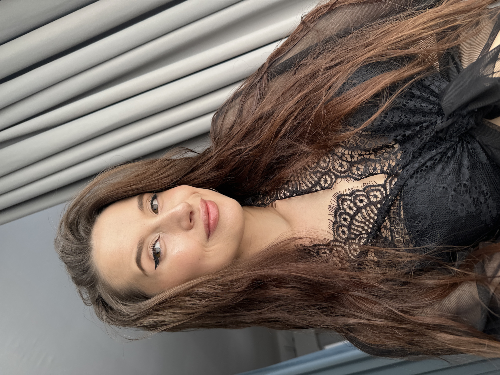 A portrait-style photograph features a young woman with long, flowing dark brown hair, gently parted and cascading over her shoulders and chest. She has fair skin, brown eyes accentuated with winged eyeliner and mascara, and well-groomed eyebrows. Her lips are a natural pink, slightly parted in a soft, welcoming smile as she looks directly at the camera. She appears to be wearing a delicate black top or lingerie piece made of intricate lace and sheer mesh fabric, visible around her neckline and décolletage.
The background consists of light gray horizontal slats or blinds on the upper portion, transitioning to a solid gray wall below. A dark blue or gray surface is also visible in the lower right corner, possibly furniture. The lighting is soft and even, suggesting an indoor setting during daytime. The woman's expression conveys calmness and approachability. The overall image captures her posing for the camera. There is no visible text in the image. The location context for this image is Yekaterinburg, Russia.