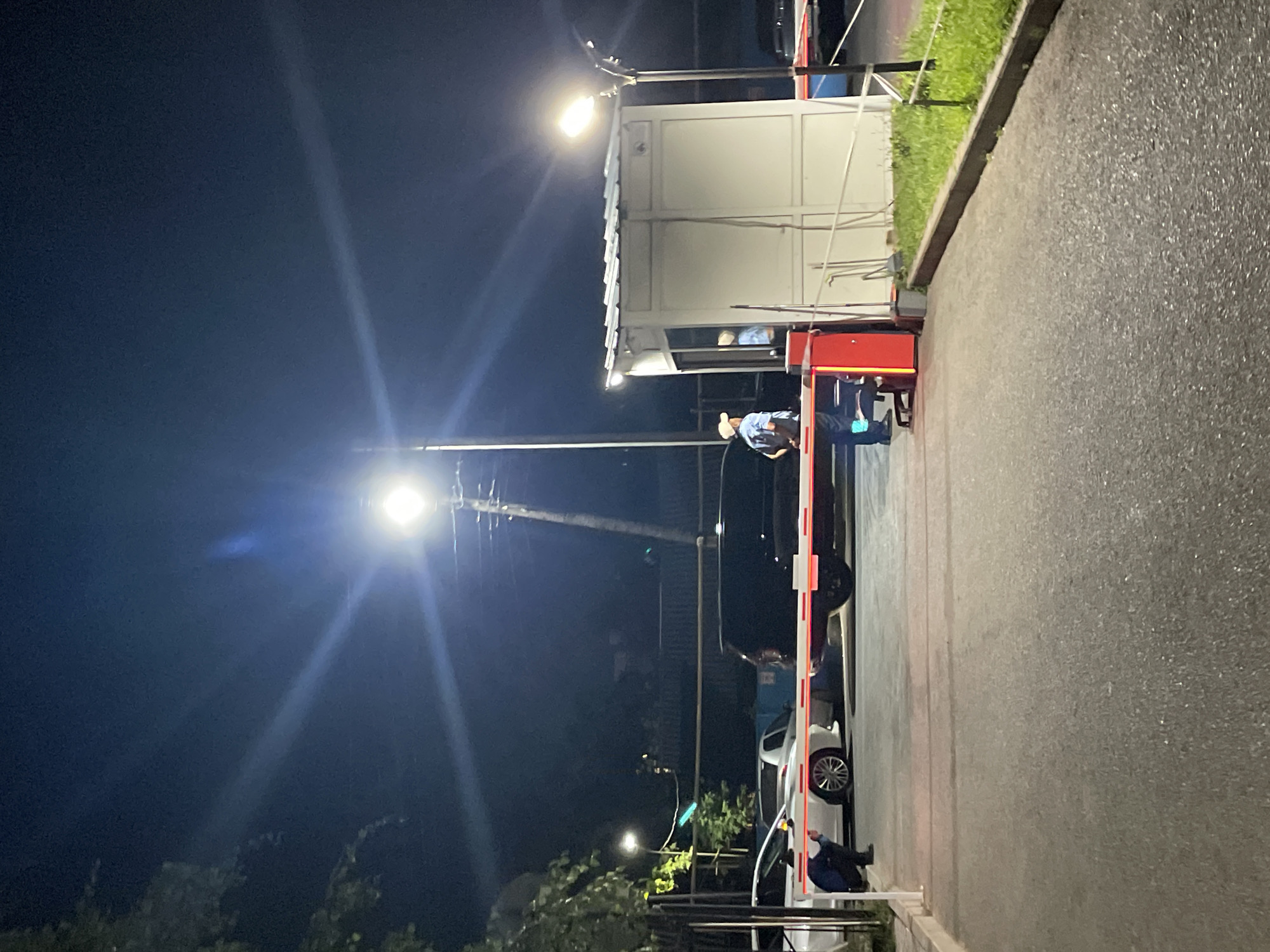 An outdoor, night-time scene at a parking lot entrance in Almaty, Kazakhstan, shows a security guard in a light blue uniform, wearing a cap and carrying a backpack, standing near a white booth with a metallic roof. The guard is facing a red and white striped barrier arm that is lowered. Another individual, possibly another guard, is visible inside the booth. Several cars are parked behind the barrier, including a white sedan and a black van. The area is illuminated by streetlights, creating lens flares in the image. The ground is paved, and there is a strip of grass along the right side of the frame.