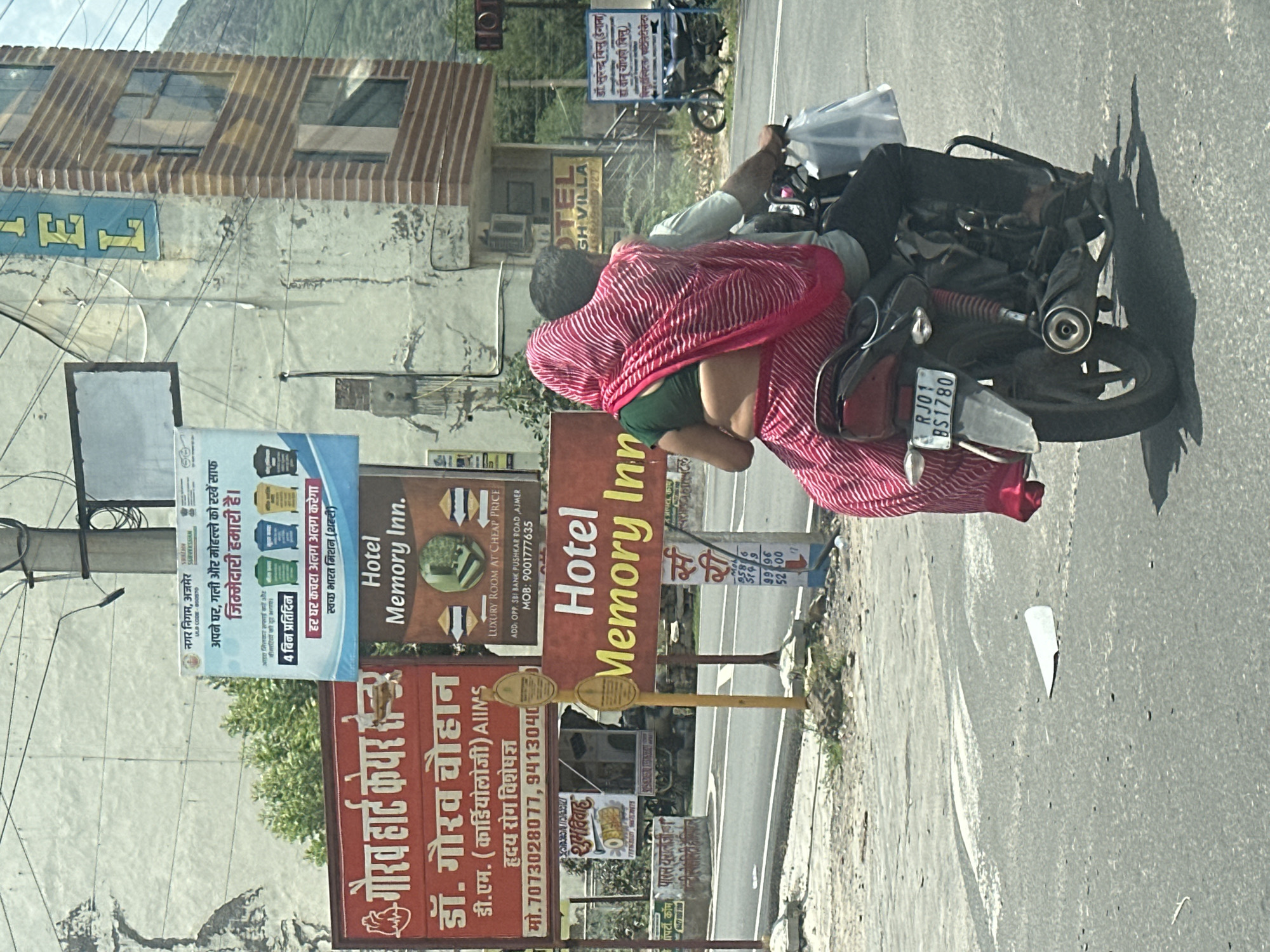 This is a medium shot, taken from a vehicle, of a couple riding on a motorcycle in Ajmer, India. The couple is dressed in traditional Indian clothing, with the woman wearing a pink and white striped sari. The motorcycle has a license plate that reads "RJ01 BS1780". In the background, there are several signs, including one for "Hotel Memory Inn." There is also a building with "HOTEL" written on it vertically in large blue letters. The environment appears to be a city street on a sunny day, with some vegetation and mountains visible in the distance.