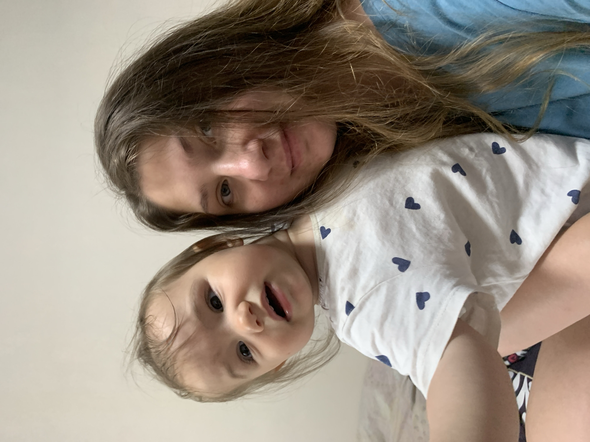 A close-up, selfie-style photograph captures a woman and a young child, likely her daughter, in an indoor setting. The woman, with long, light brown hair, is positioned on the right side of the frame, looking towards the camera with a soft smile. Her daughter, with dark hair and wide eyes, is nestled into her side on the left, her face turned slightly away from the camera, conveying a curious or perhaps slightly surprised expression. Both are casually dressed; the child wears a white t-shirt adorned with small dark blue hearts, and the woman is in a blue top. The background is a plain, light-colored wall, suggesting a domestic environment, possibly a bedroom, given the hint of bedding on the lower left of the image. The lighting is soft and natural, highlighting their faces. No specific landmarks or elements that could pinpoint a location within Chaikovsky, Russia, are visible in the image.