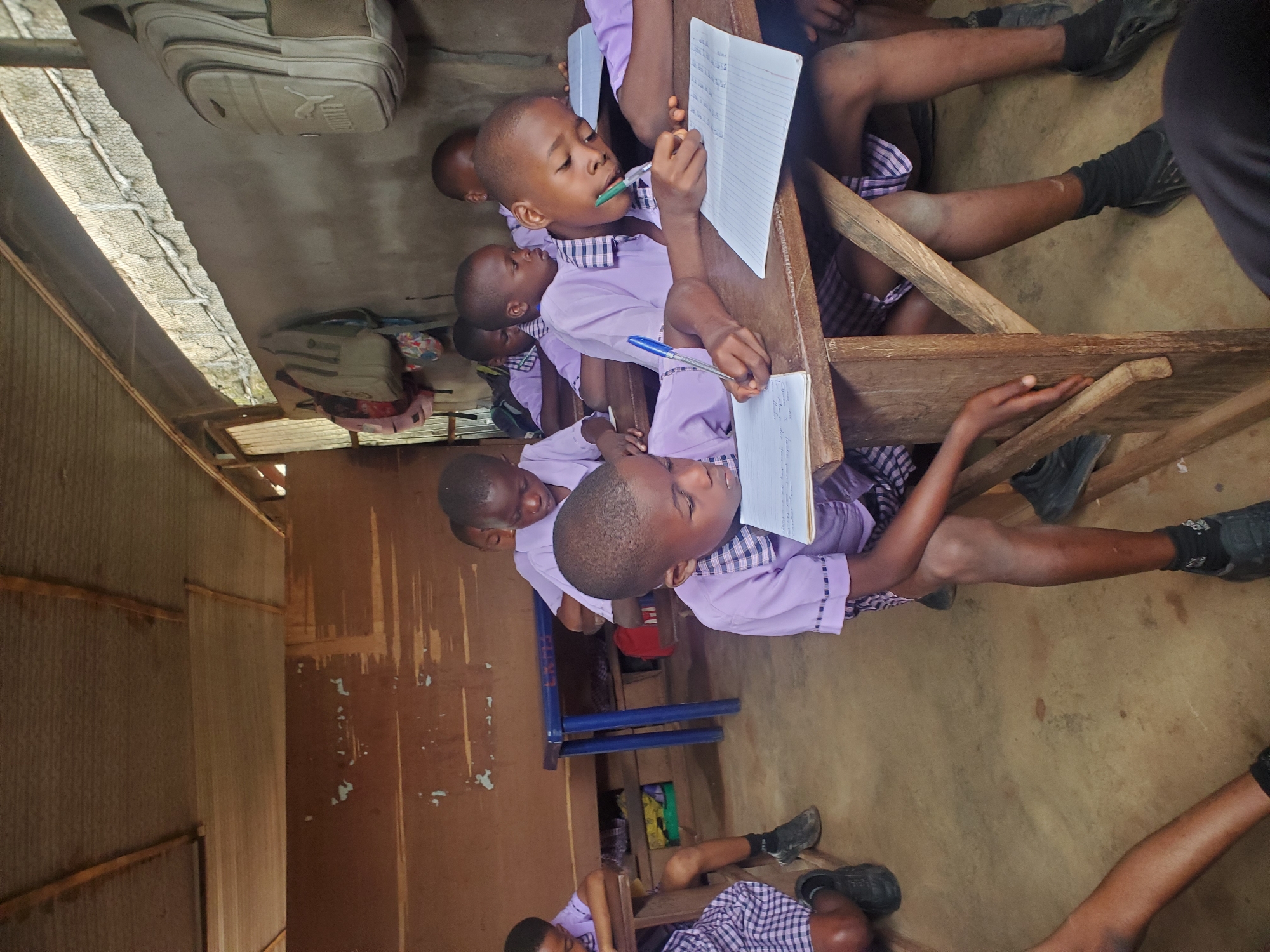 The scene depicts an elementary school classroom in Ukwu-Abwa, Nigeria. Several young students, wearing light purple uniforms with checkered collars and sleeves, are seated at wooden desks. The children are writing on notebooks with pens, focusing intently on their work. The classroom appears sparsely furnished, with visible corrugated metal walls and a simple dirt floor. A few backpacks are visible.