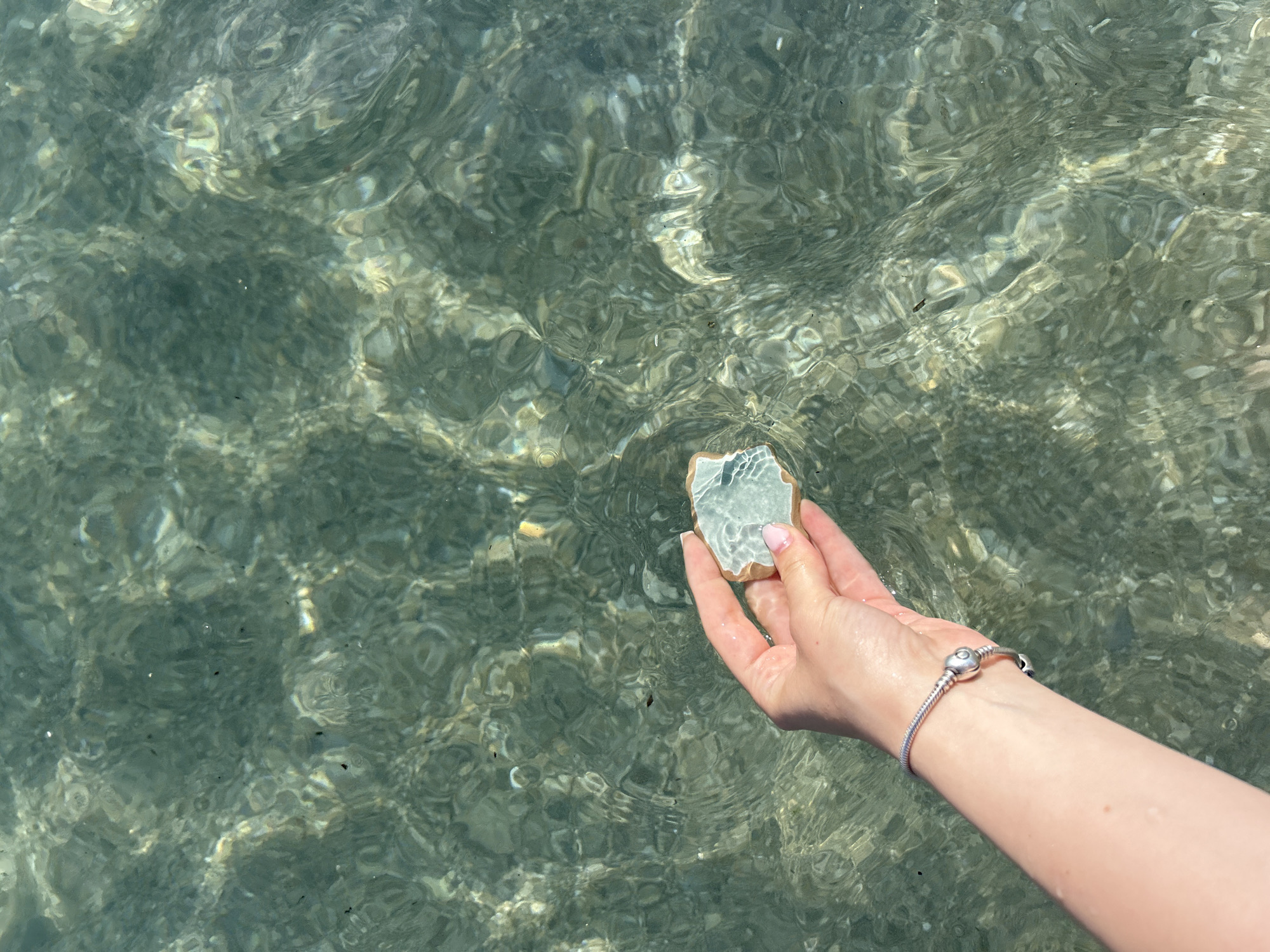 A person is holding a stone in their hand above a body of water. The water is clear and slightly rippled, with the sunlight creating patterns on the surface. The stone is light-colored and appears to be a piece of ceramic with a glaze. The person's hand is fair-skinned and has a silver bracelet on their wrist. The location appears to be a shoreline where the water is shallow enough to see the bottom. The city is Unknown, Unknown.