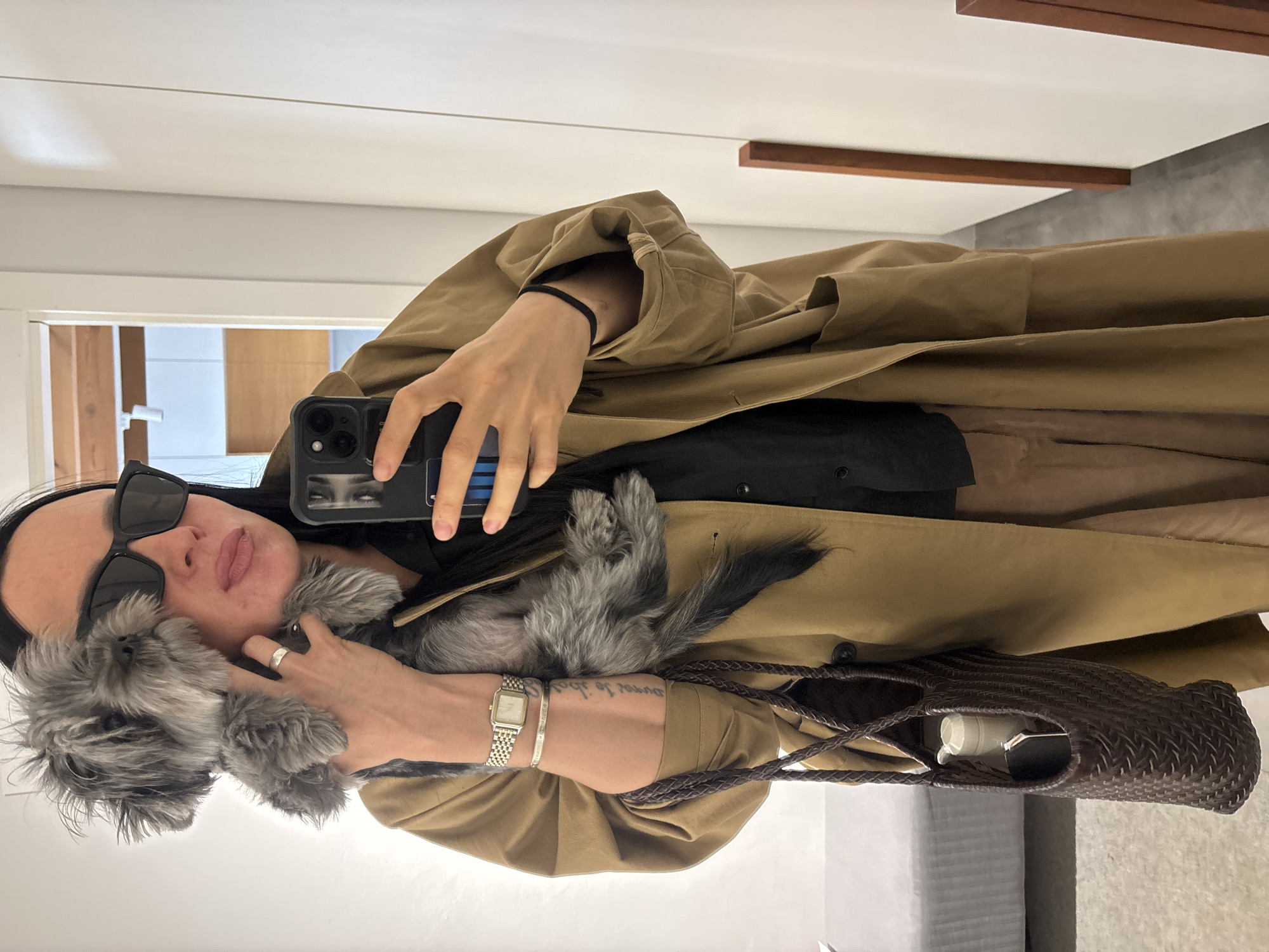 An individual, appearing to be female, is captured taking a mirror selfie indoors. She wears a tan or khaki trench coat over a black undershirt, paired with large black sunglasses. Her dark hair is styled back from her face. She holds a small, fluffy gray dog close to her left cheek. The dog has dark eyes and a black nose.
On her left hand, she wears a silver ring, and on her left wrist, a silver watch with a square face and a link bracelet. Below the watch, another silver bracelet is visible, bearing the French inscription "j'aime ta folie" (I love your madness). A tattoo on her inner forearm begins with "je t'aime j'ai vu..." (I love you I saw...). She uses her right hand to hold up a black smartphone, equipped with multiple camera lenses, to take the photo through the mirror.
A braided dark brown bag hangs from her left arm, and its contents appear to include a white bottle or container and another dark object. The background reveals a brightly lit interior space featuring white walls and ceiling, with visible horizontal wooden beams or accents. The floor appears to be covered with a light-colored rug or carpet. The setting is a modern indoor environment, consistent with a casual mirror selfie. The provided context suggests the location is Chaikovsky, Russia. The time of day is not explicitly indicated by the indoor lighting.