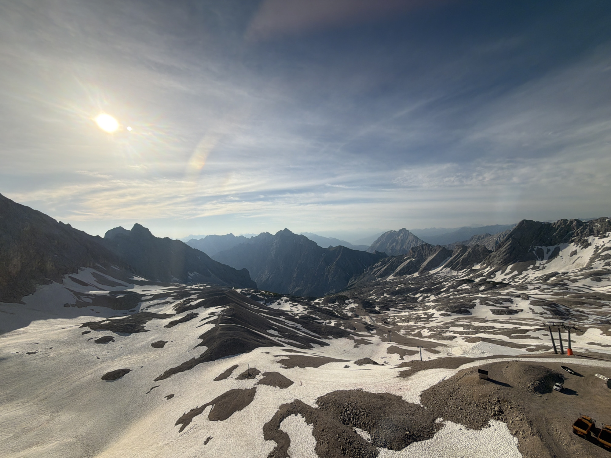 This is an aerial, wide-angle, panoramic view of a mountainous landscape near Garmisch-Partenkirchen, Germany. The scene is dominated by rugged, snow-dusted peaks and a vast expanse of terrain characterized by patchy snow and exposed dark earth and rocks. In the foreground, the snow cover is broken, revealing rocky areas and what appears to be a ski slope or a pathway. Several ski lift pylons are visible, suggesting a ski resort. There are a few small vehicles, including a white car and what appear to be construction or maintenance vehicles at the lower right. A single person is visible in the distance on the snow-covered slope. The sky is a pale blue with wispy, cirrus clouds. The sun is shining brightly in the upper left quadrant, casting a strong glare and lens flare across the image. The overall impression is one of a high-altitude, alpine environment, possibly during the late spring or early summer when snow is still present but melting.