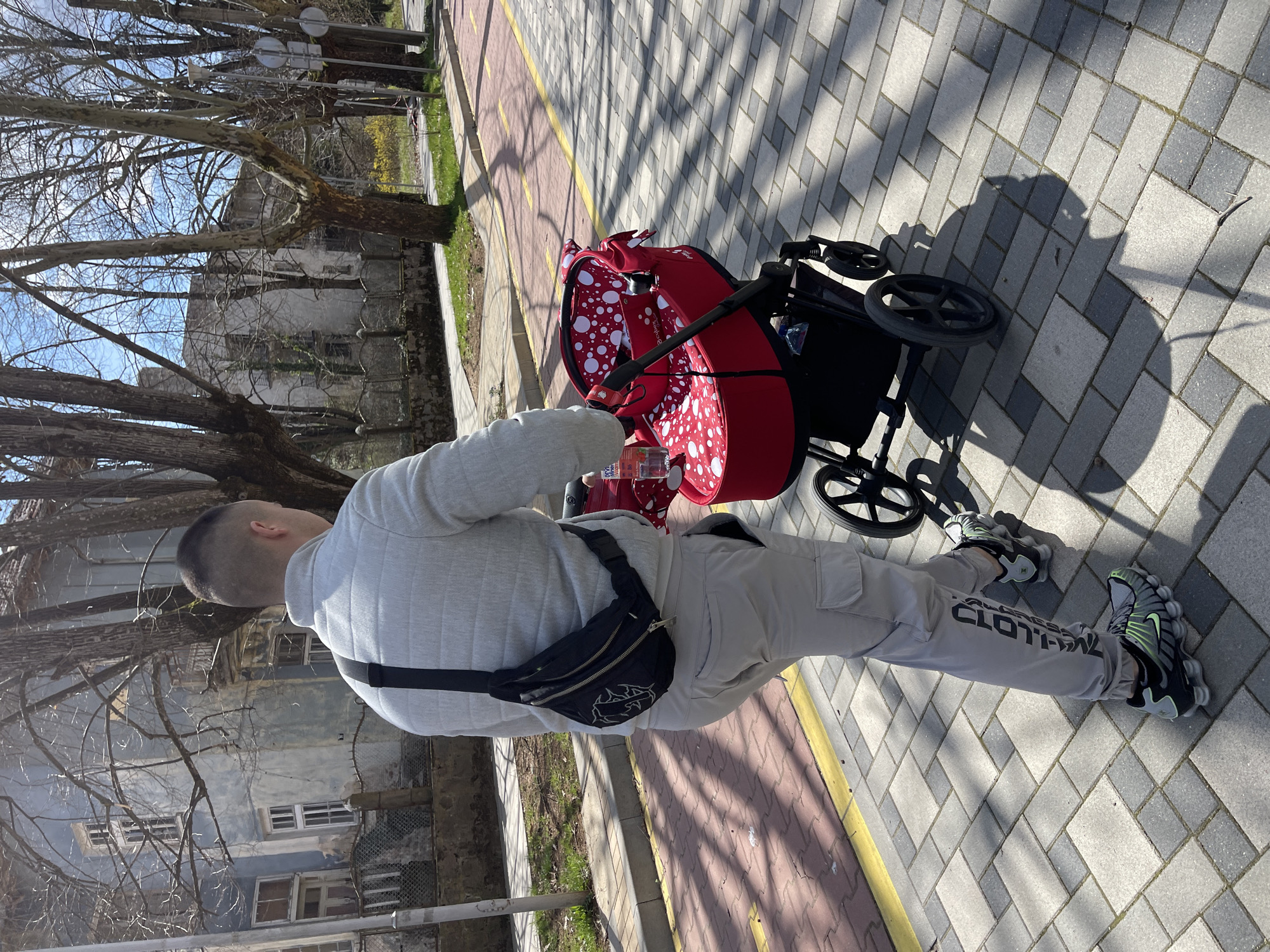 A medium-shot presents a man wearing a light gray, quilted jacket, beige cargo pants with text on the lower leg, a black fanny pack, and black and green sneakers, pushing a red baby stroller with white polka dots on a brick sidewalk in Varshets, Bulgaria. The sidewalk has a yellow line marking its edge. Bare tree branches are visible on the left. A weathered building with visible windows and a wire fence runs along the left side of the sidewalk. The scene appears to be during daytime, casting shadows.