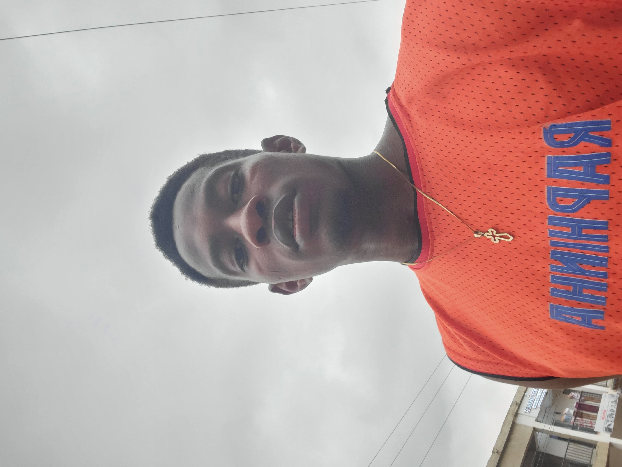 A close-up, waist-up shot captures a young Black man in Egbagu, Nigeria, under an overcast sky. He is wearing an orange sleeveless jersey with a black trim around the neck and armholes. A gold chain with a cross pendant rests on his chest, visible against the jersey. The jersey has a blue graphic design across the front. In the background to the left, a building with signage and balconies is partially visible. The lighting suggests it is daytime, likely late morning or afternoon. The man is looking directly at the camera with a neutral expression.