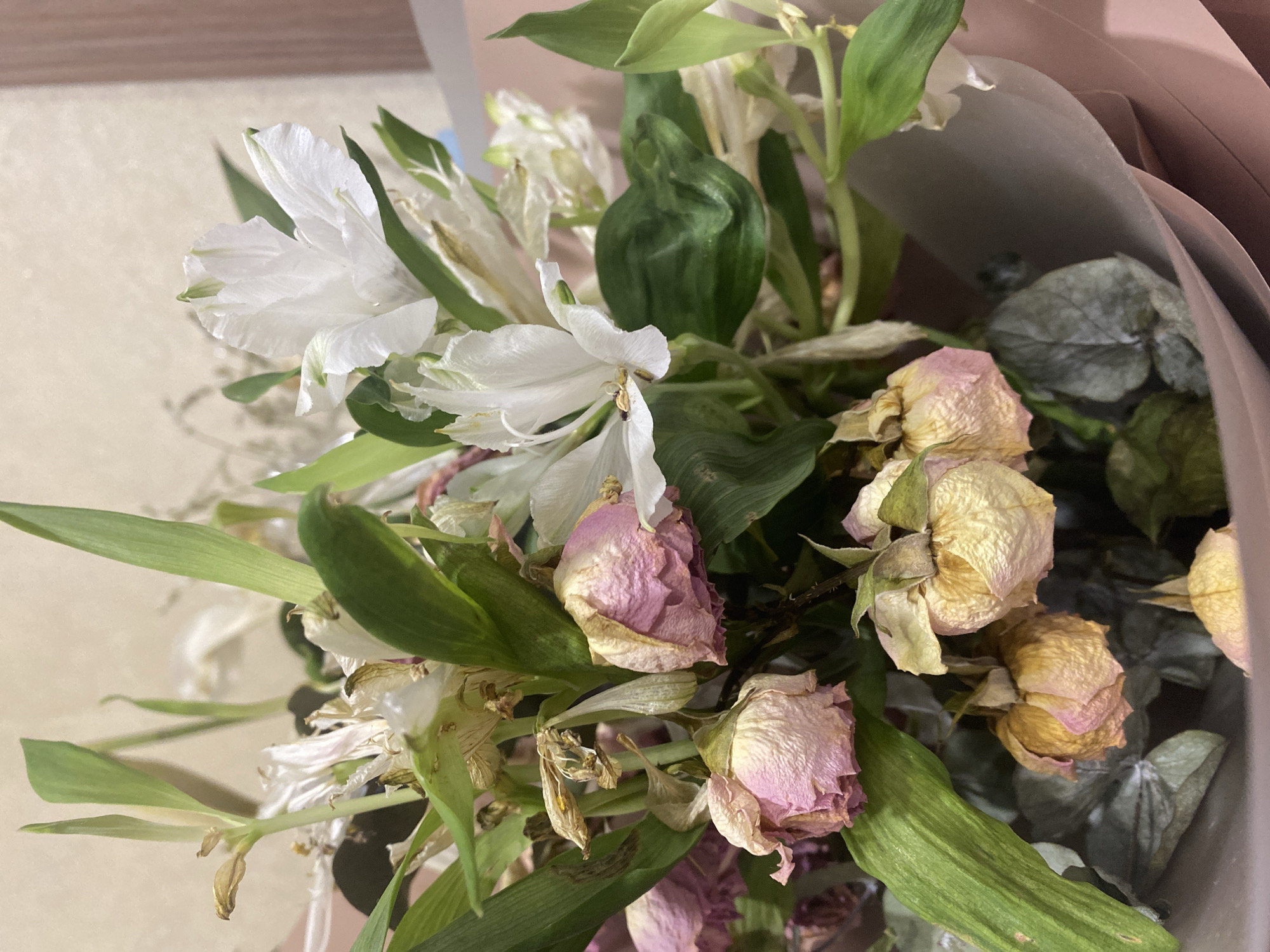 A bouquet of flowers, some fresh and white, others wilted and pink and yellow, is presented. The bouquet is wrapped in pink paper. The background is a neutral, textured wall. The image appears to be a close-up shot taken indoors, possibly in a domestic setting. There are no people or discernible actions depicted. The focus is solely on the floral arrangement.