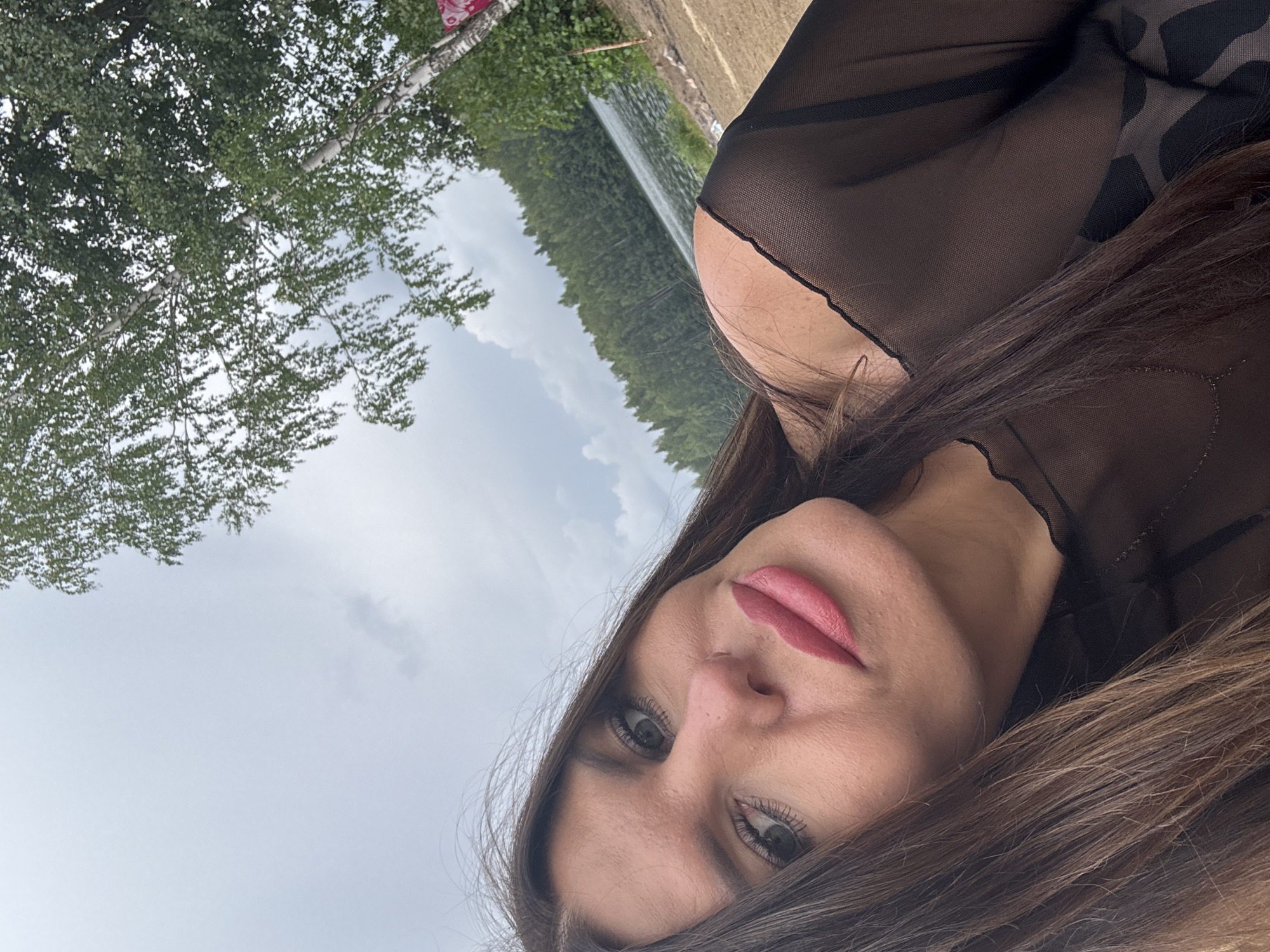 This is a selfie taken outdoors in Gol'yany, Russia. The photo is a close-up of a woman's face and upper body. She has long, brown hair and is wearing a sheer black top. In the background, a lake is visible, bordered by a dense forest of trees. The sky is overcast.