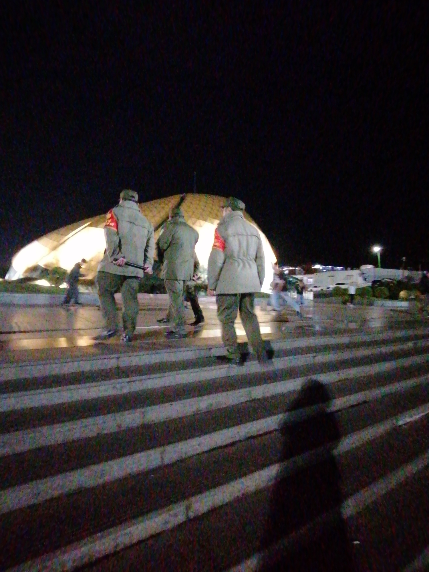 In the city of Da Lat, Vietnam, at night, a group of individuals in military-style uniforms are walking on a crosswalk. They are illuminated by artificial lights, and a large, modern building with a curved, illuminated roof is visible in the background. The individuals are wearing what appear to be olive-green uniforms with red armbands. One person in the foreground is holding a black baton. The crosswalk is wide and appears to be made of striped concrete or asphalt. The overall scene suggests a security or patrol detail in a public area.
