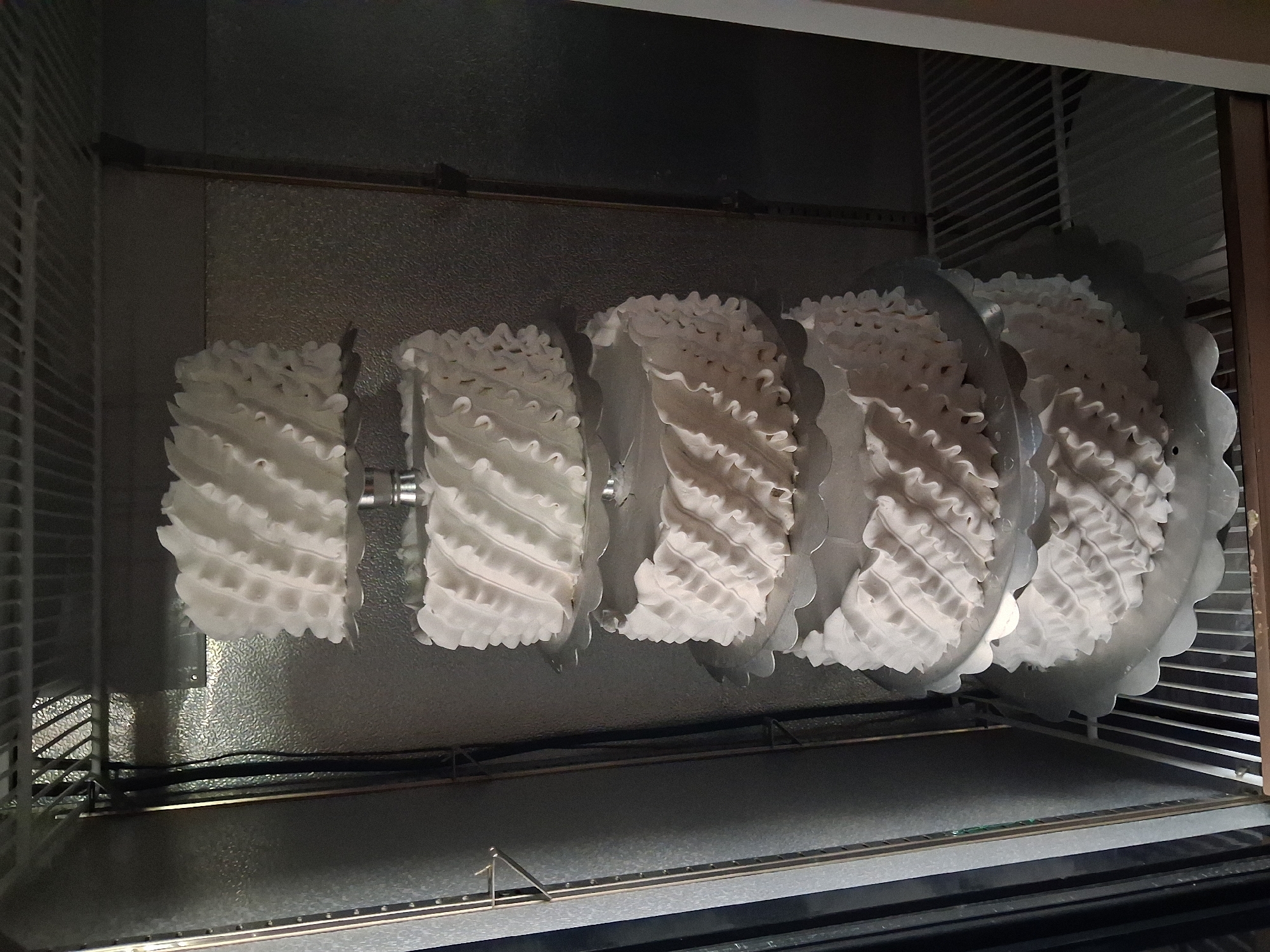 A five-tiered cake decorated with white frosting ruffles is displayed inside a commercial refrigerator. The cake layers are stacked on silver scalloped cake stands. The refrigerator has a textured metallic interior and wire shelves. The location is in the city of Guldala, Kazakhstan.