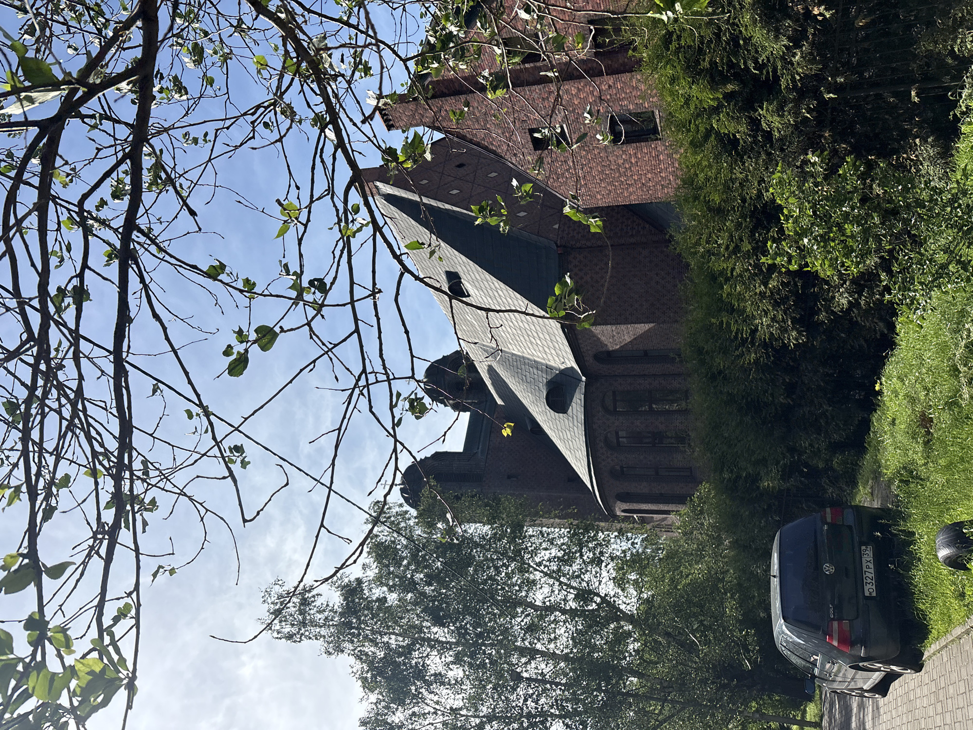 The image, captured in Kaliningrad, Russia, depicts a building with a dark brick facade and a distinctive, angular roofline. The building appears to be old, with arched windows visible. In front of the building, a dark-colored Volkswagen Golf is parked on a paved surface next to a grassy area. Lush green trees and foliage surround the scene, with a prominent tree in the foreground partially obscuring the view of the building. The sky is a clear blue with some wispy clouds, suggesting it is daytime with fair weather. The overall impression is a quiet, perhaps residential, setting. The license plate on the car is visible and reads "O 327 PX 39". No people are visible in the image.