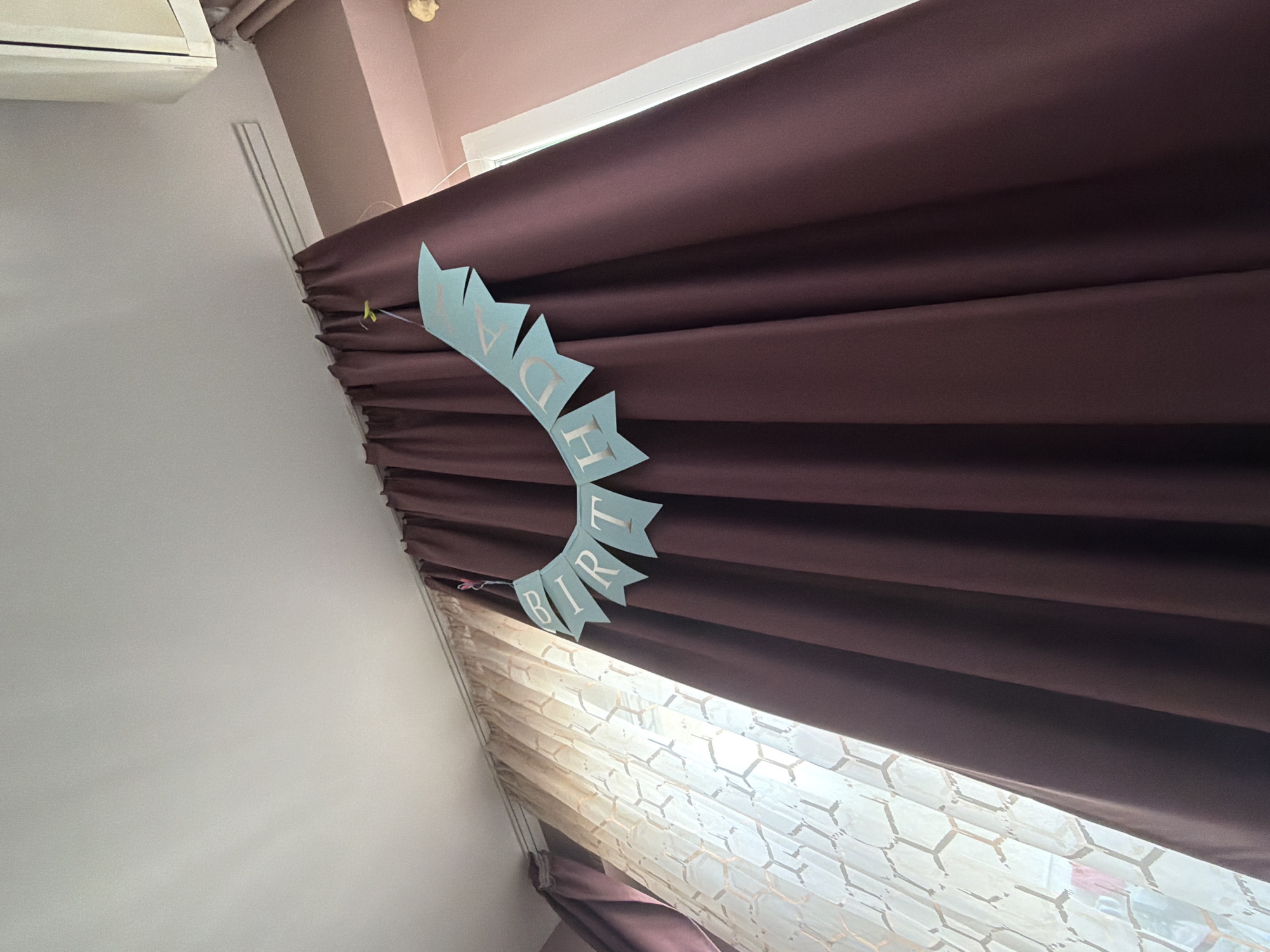 A rotated, indoor, medium shot captures a scene in what appears to be a room in Sofia, Bulgaria. A section of the wall is visible, along with a window covered by brown curtains and a sheer honeycomb-patterned blind. A light blue "Birthday" banner is attached to the curtain. The color palette of the scene consists of light brown, white, and a shade of brown-purple in the curtains. The artificial light source illuminates the blind behind the dark curtains. A white air conditioner is visible in the upper left corner. There are no people in the shot.